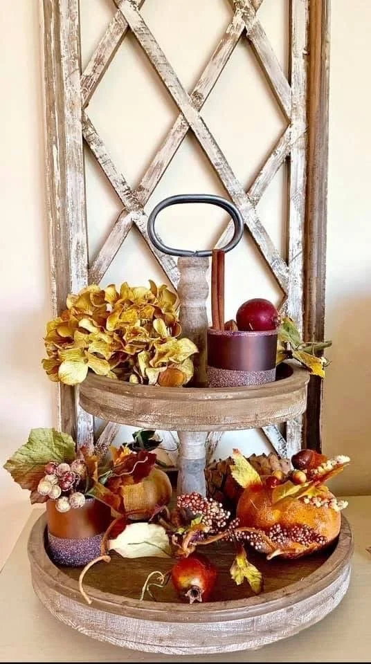 A two-tier wooden display stand with autumn-themed decorations, including artificial pumpkins, leaves, berries, and a bouquet of yellow flowers on an oxidized holder, set against a rustic crosshatched wooden backdrop.