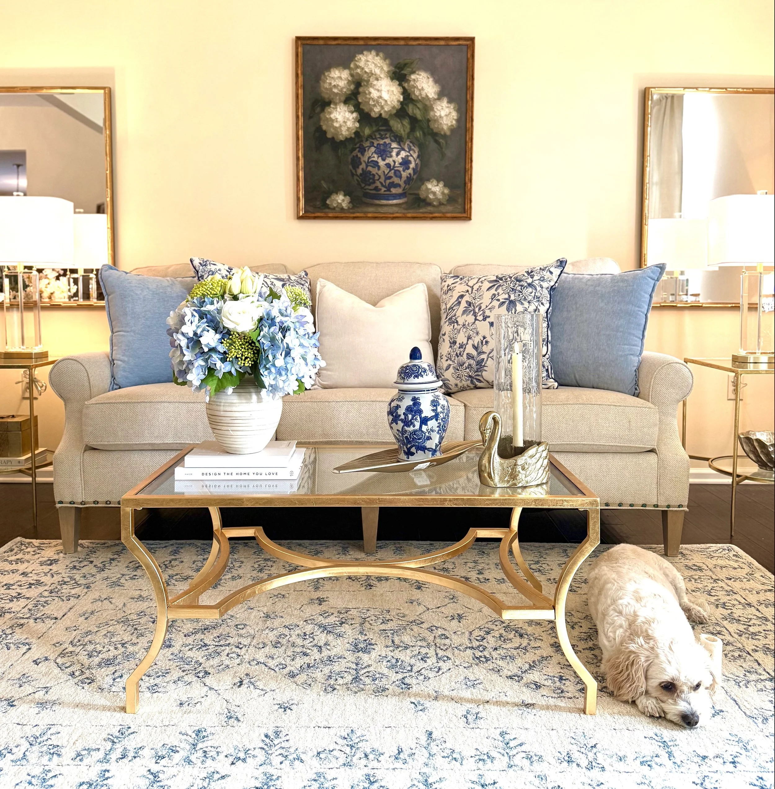 A thoughtfully styled living room featuring a neutral sofa layered with blue and white patterned and solid pillows, flanked by matching crystal and gold table lamps on gold and glass end tables and gold mirrors behind each lamp. Gold framed artwork s