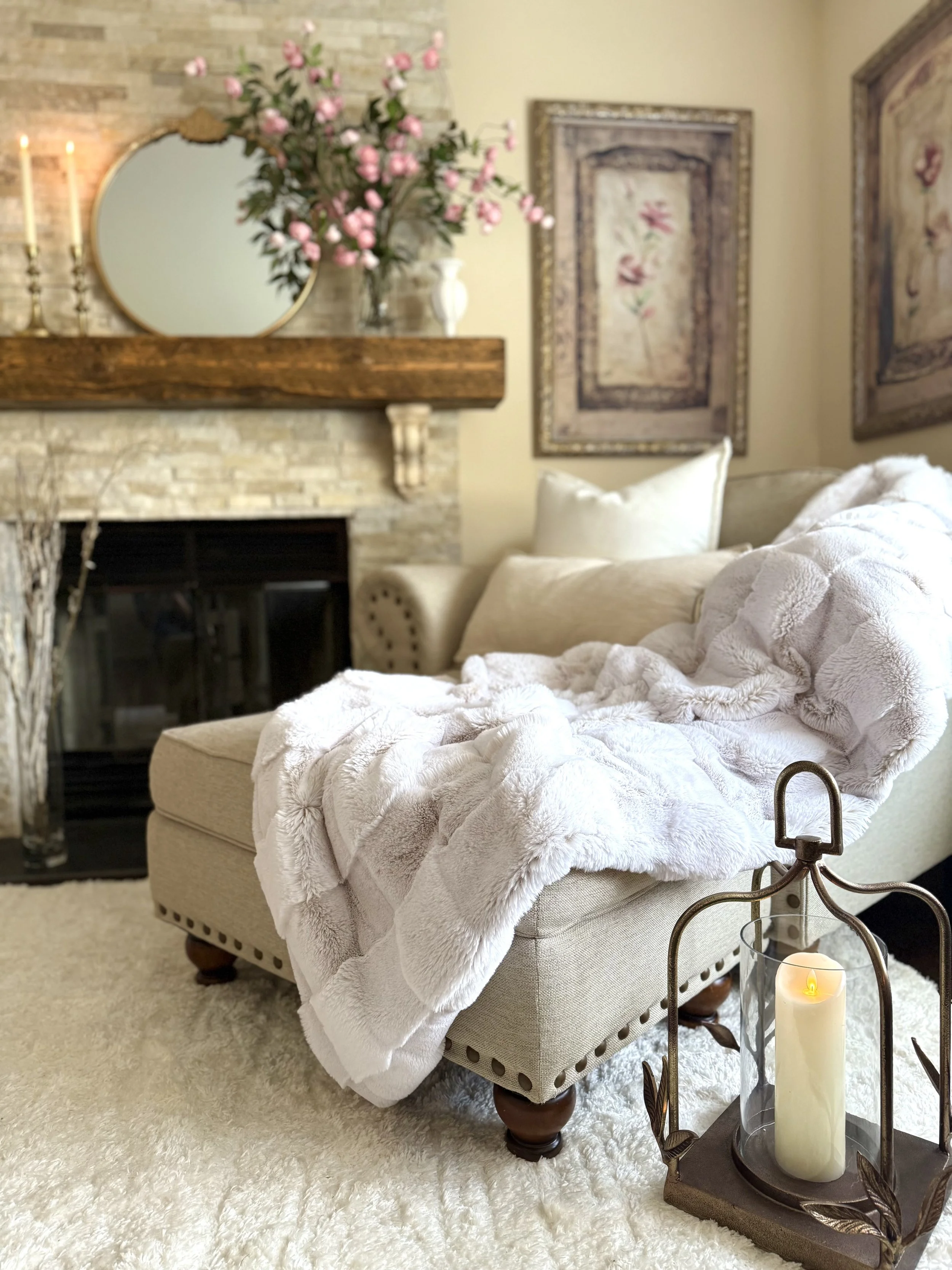 Soft neutral living room featuring a cozy upholstered chaise, layered textiles, framed pink artwork, a styled ledgerstone fireplace with a rustic wooden mantle held up by antiqued corbels with large decorative pink florals, a white vase, a shag white