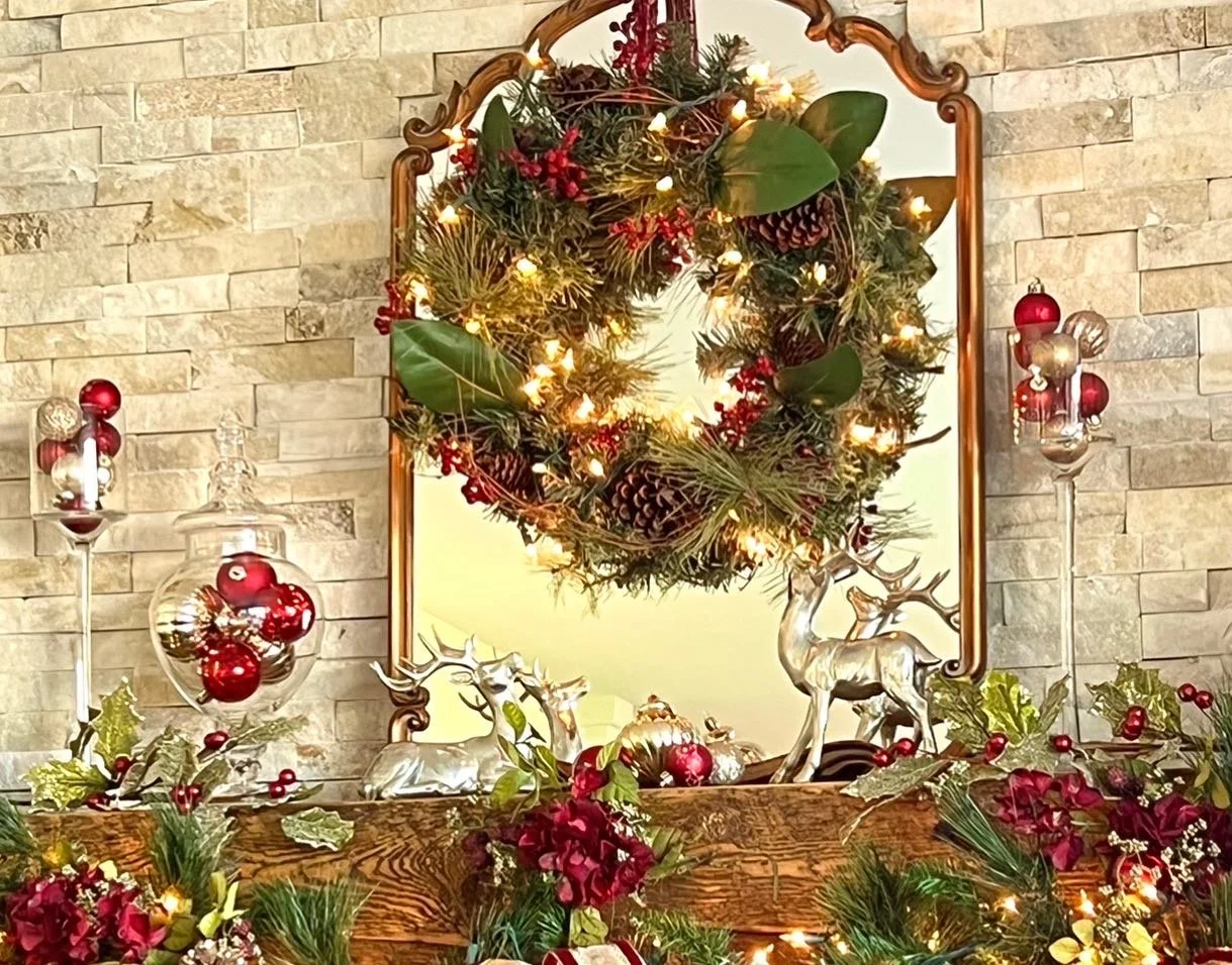 Decorative Christmas wreath with pinecones, red berries, greenery, and fairy lights hangs above a fireplace mantel. The mantel is decorated with red and gold ornaments, holly, and greenery, along with a silver reindeer figurine. The background featur