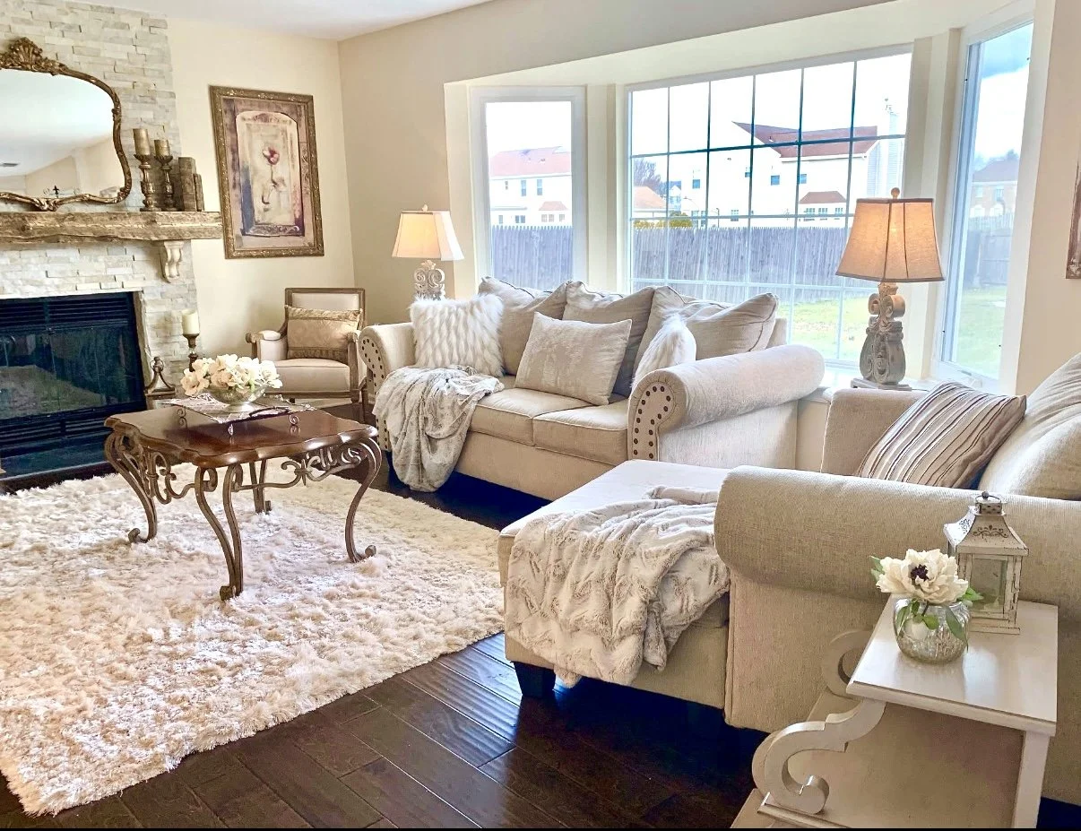 A cozy living room with cream-colored sofas, a wooden coffee table, a stone fireplace, and large windows providing a view of neighboring houses. The room is decorated with lamps, pillows, a floral arrangement, and soft throws on the furniture.