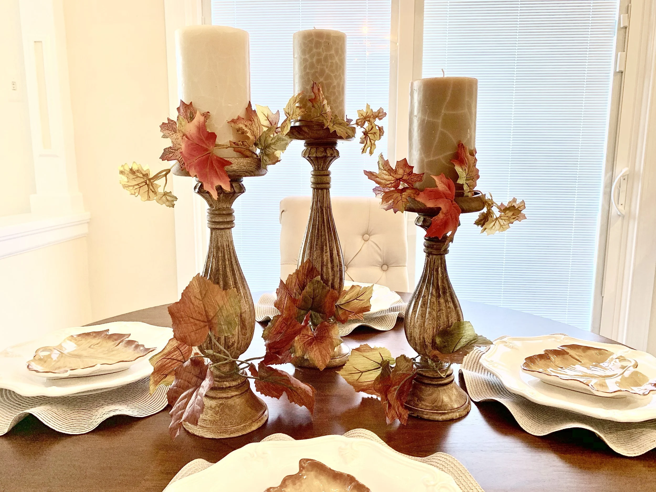 Autumn-themed dining table centerpiece with three tall candles, artificial fall leaves, and decorative plates.