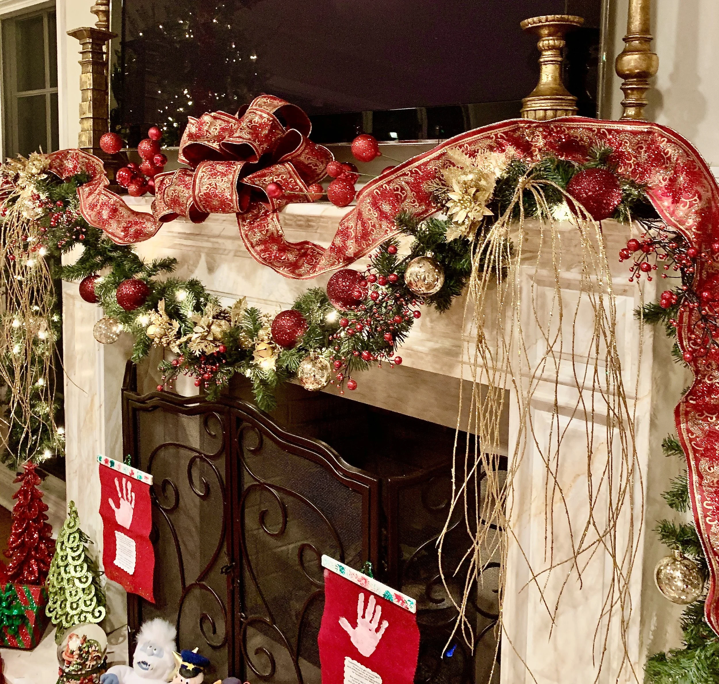 Christmas holiday fireplace mantel decorated with red, gold, and green ornaments, ribbons, and greenery, with stockings hanging and various festive decorations below.