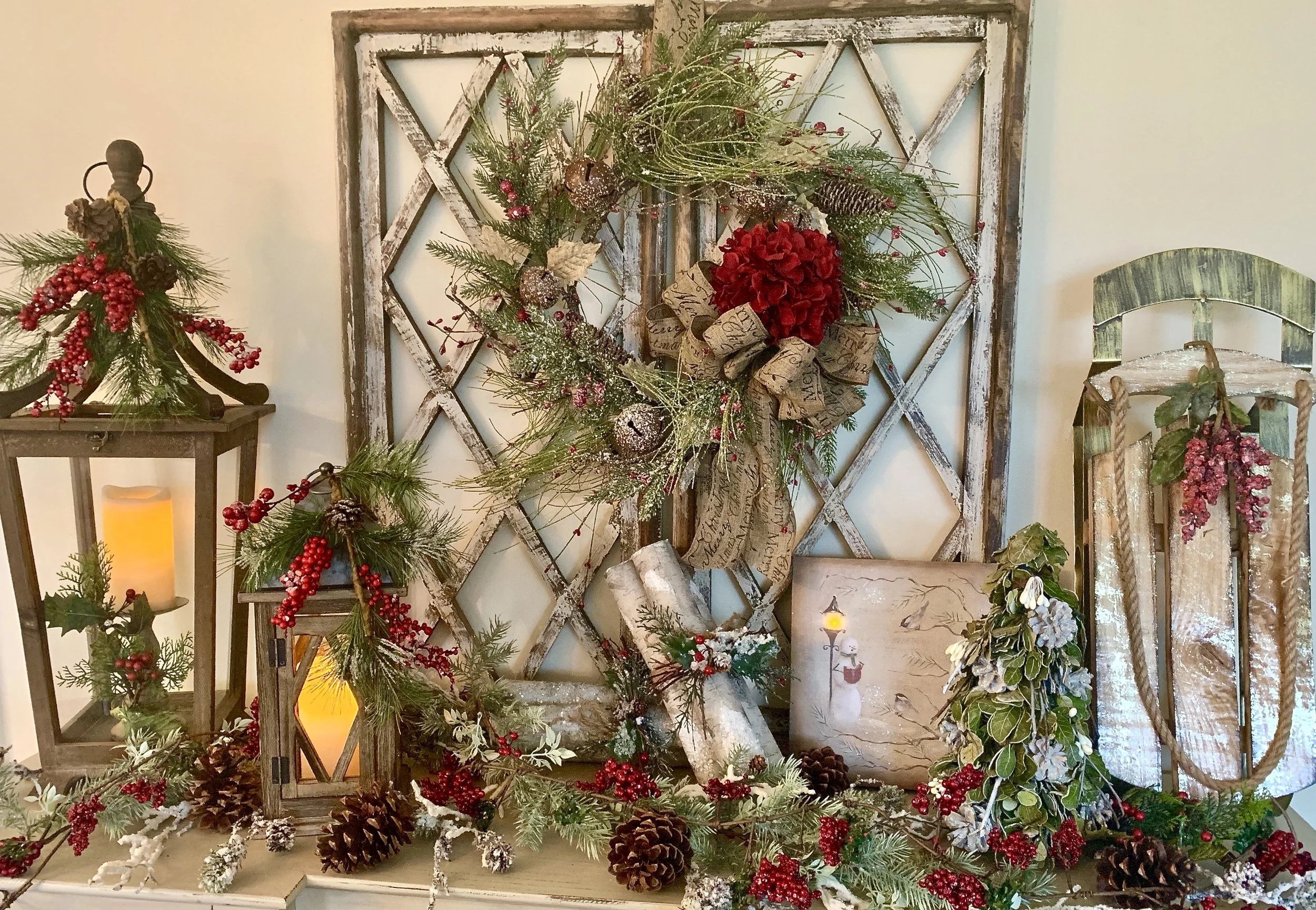 Decorative Christmas display with greenery, red berries, pinecones, lanterns, and a painting featuring a snowman and a candle with a landscape, all arranged on a table against a white wall.