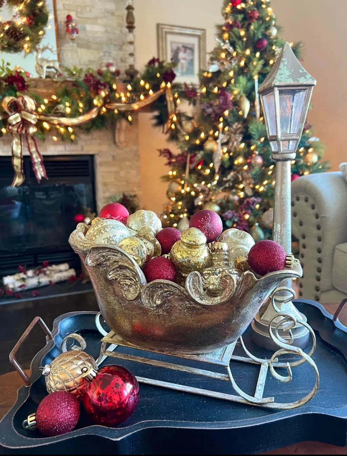 Decorative bowl filled with gold and red Christmas ornaments placed on a black tray, with a small metallic sled and a streetlamp decoration. In the background, a decorated Christmas tree with lights and ornaments, and a festive mantle with garland an