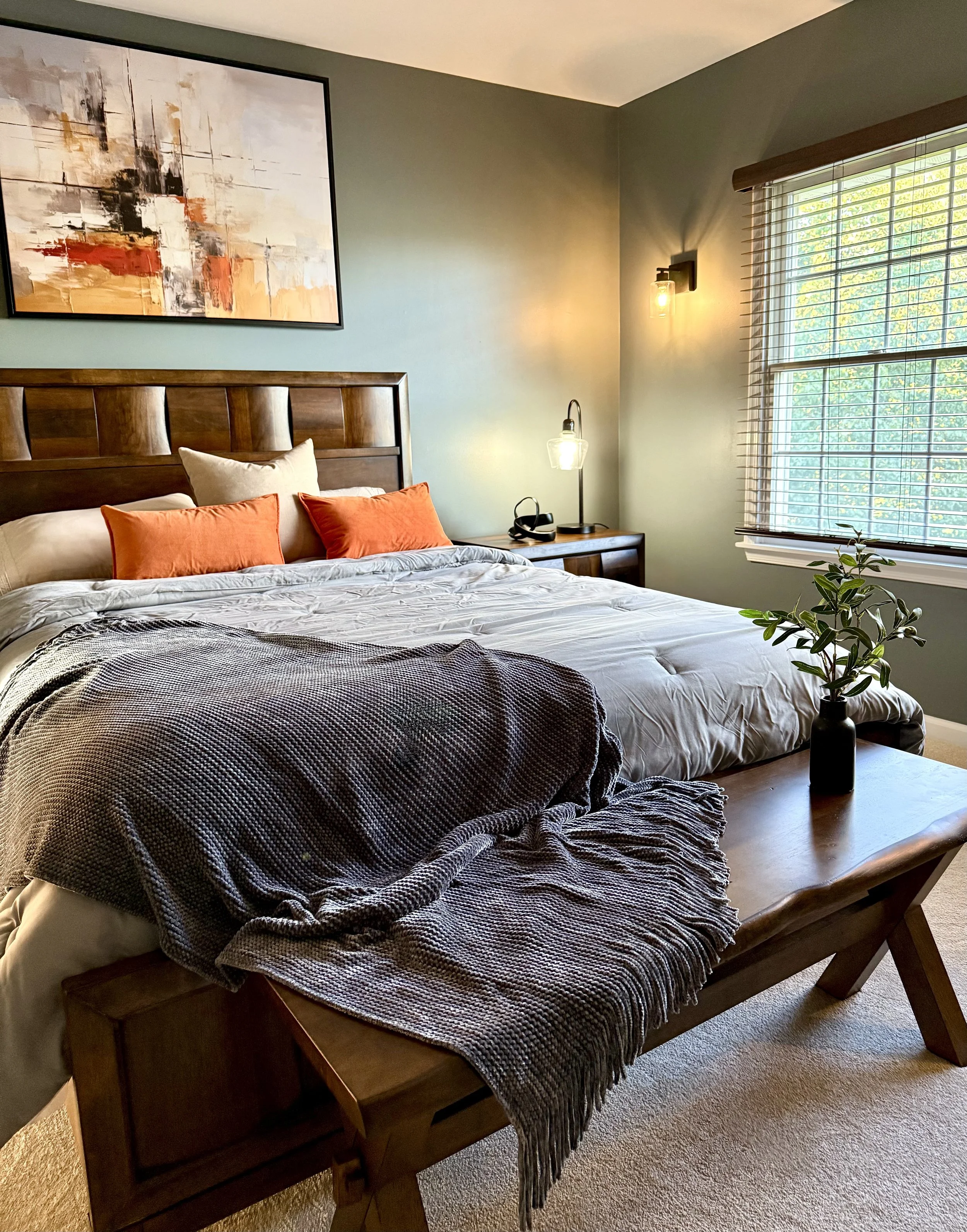 A cozy bedroom with a wooden bed frame, white bedding, orange pillows, a throw blanket, a wooden bench with a plant, a nightstand with a lamp, and a large window with blinds letting in natural light.