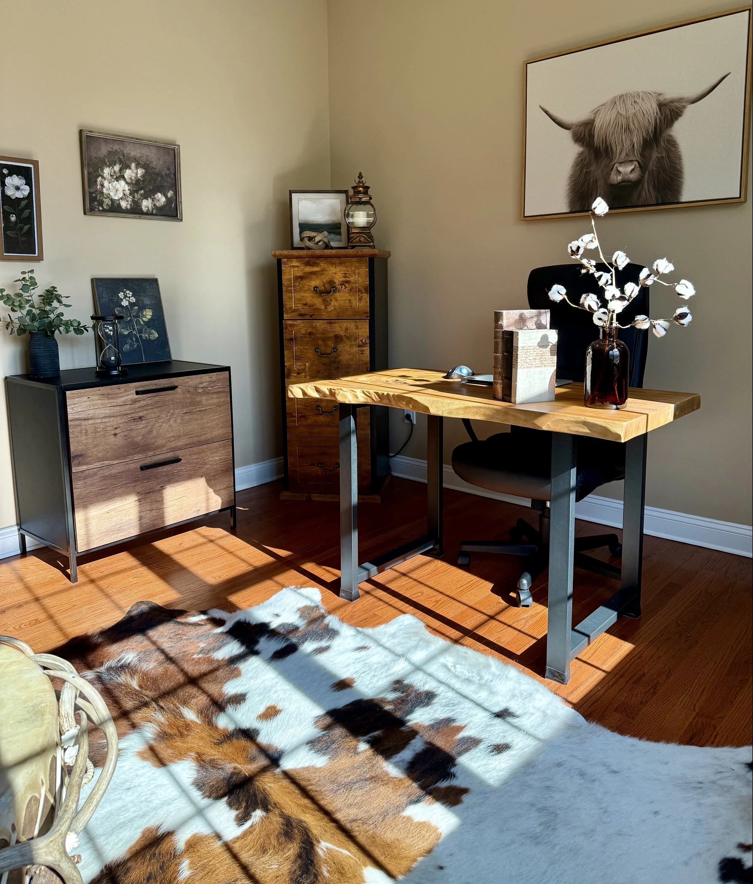 A home office with a wooden desk, black office chair, and a cowhide rug. Decor includes botanical artwork and a framed highland cow photograph on the wall, with sunlight casting shadows on the wooden floor.