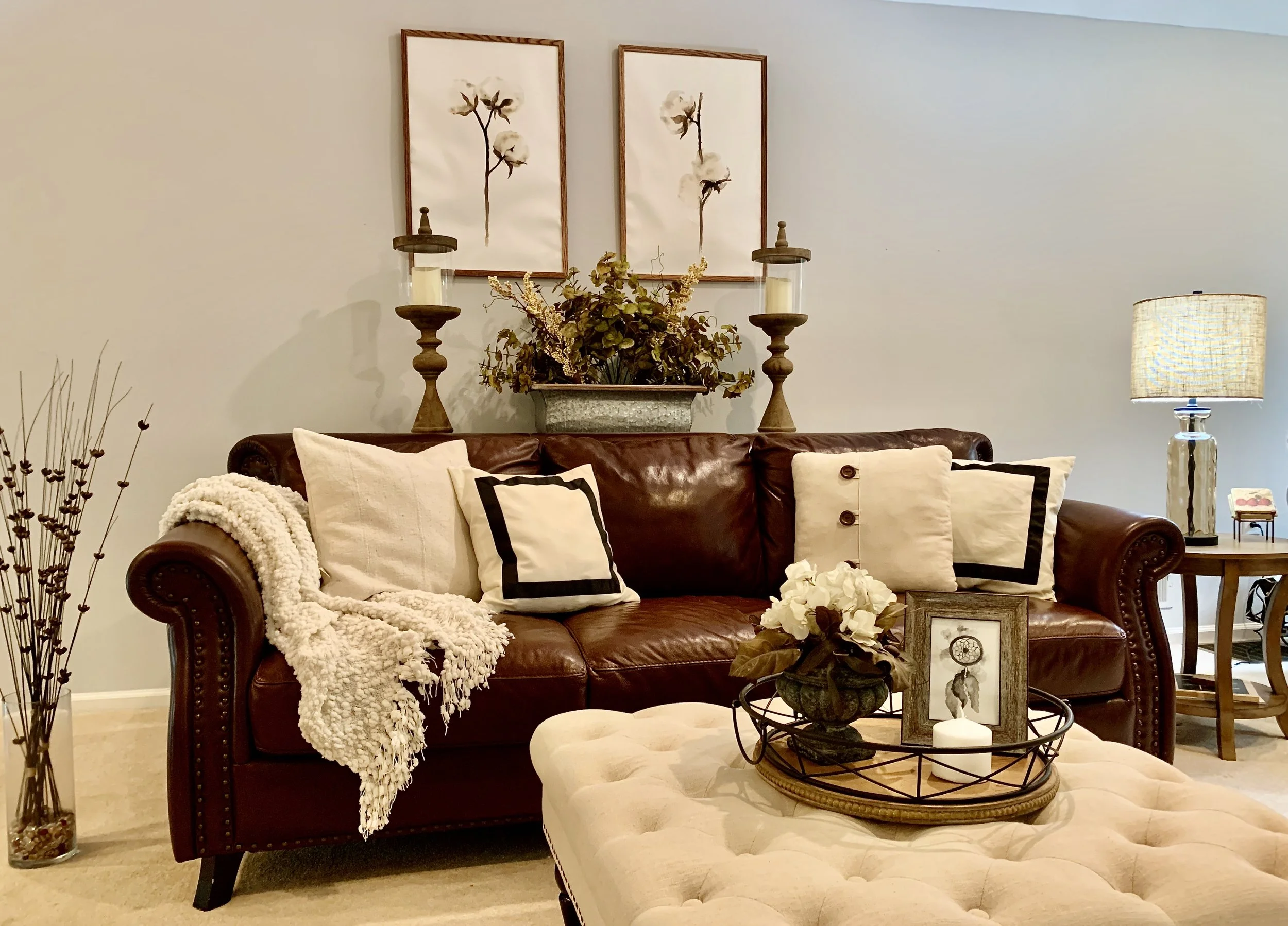 A cozy living room with a dark brown leather sofa, beige pillows, a cream throw blanket, and a tufted ottoman with a decorative tray full of flowers, framed photos, and candles. The wall behind has two framed cotton flower prints, with a planter and