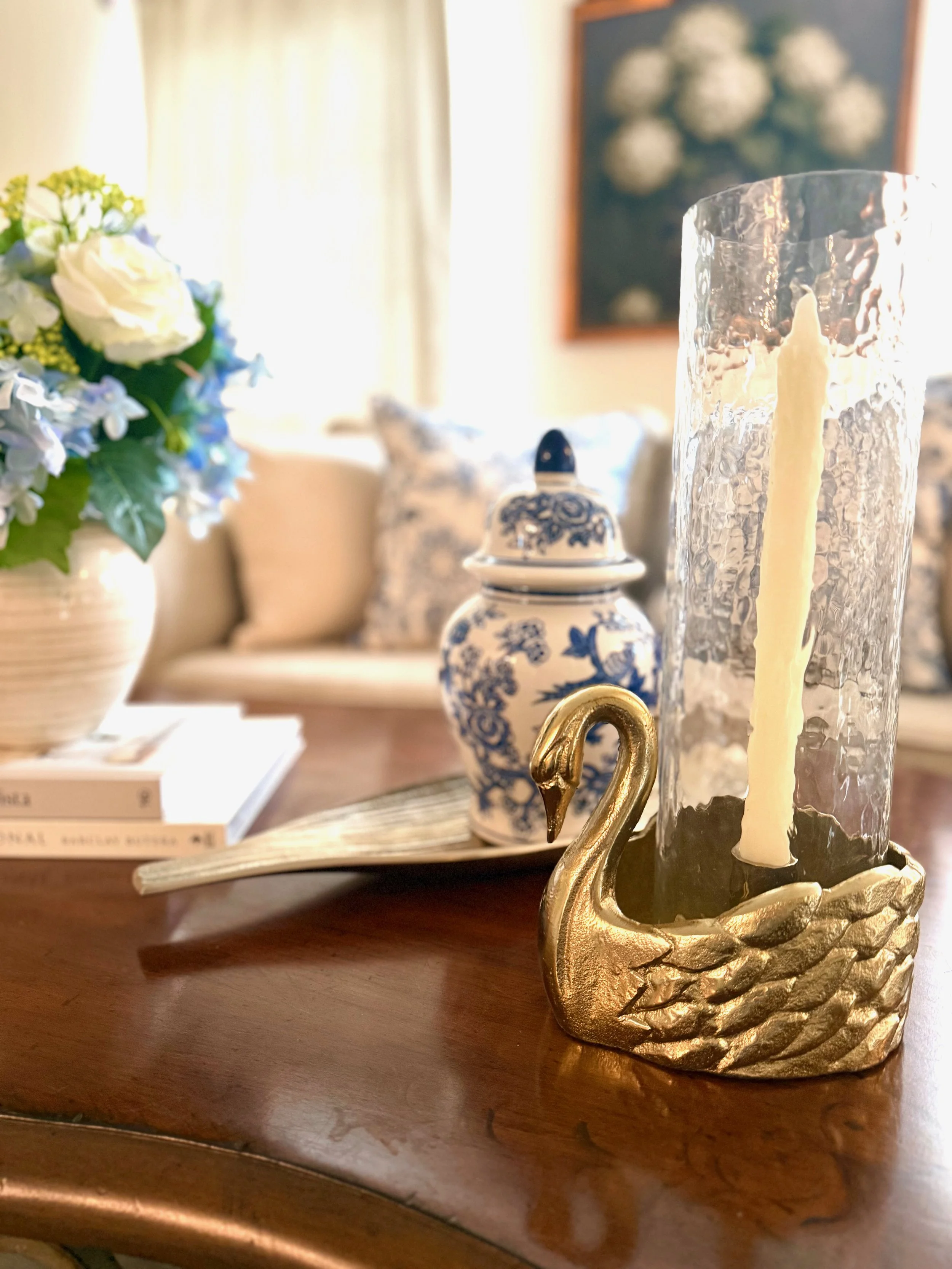 Professionally classic styled living room with a neutral beige sofa, blue and white patterned pillows, silk drapery, gold framed blue and white artwork, layered textiles, an ornate iron and wood coffee table, dark wood floors, gold swan hurricane can