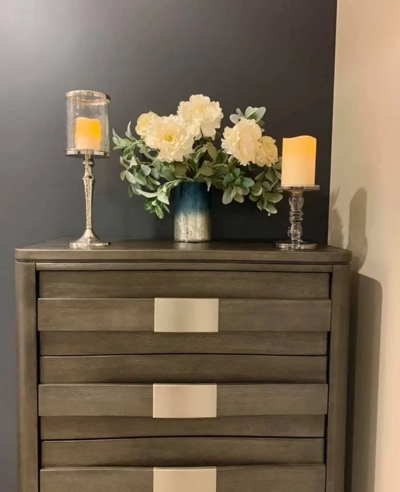A wooden dresser with four drawers, topped with a blue ceramic vase holding white flowers and greenery, flanked by two silver candle holders with candles on a dark and light wall background.