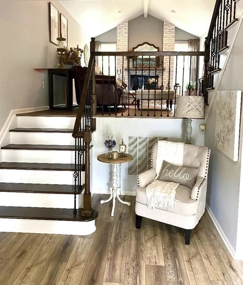 A cozy living room corner with a white tufted armchair with a pillow that says 'hello,' a small round side table with decorations, and a floor lamp with a patterned shade. An open staircase with dark wood steps and black railing leads to an upper lev