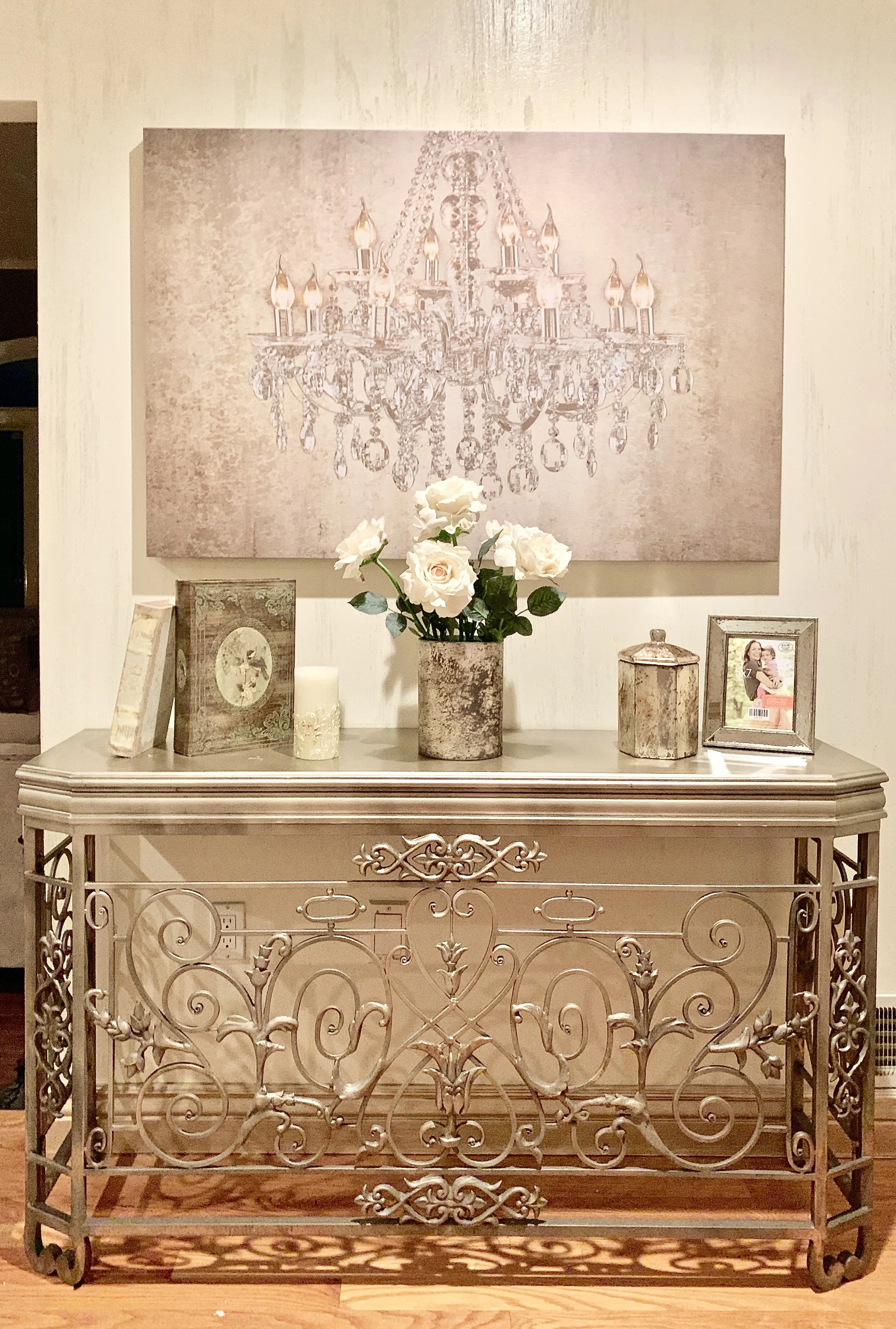 A console table with decorative items, including a vase of white roses, framed photos, and candles, in front of a large picture of a chandelier.