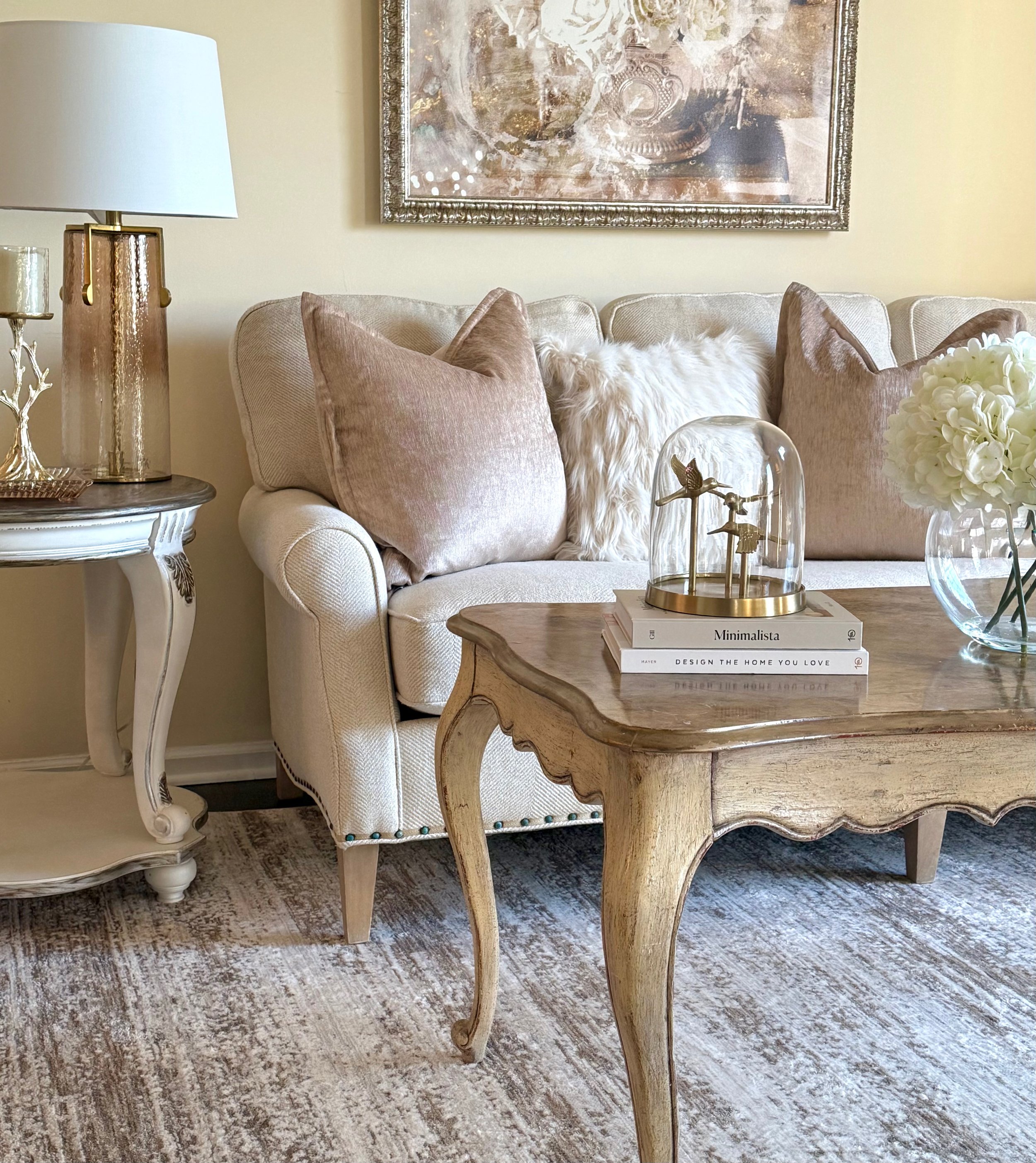Professionally decorated living room showcasing a neutral sofa, layered textiles, curved wood coffee table, gold pillows, plush fur beige and white textured pillows, framed white and gold and silver artwork, brown and beige area rug, white florals, g