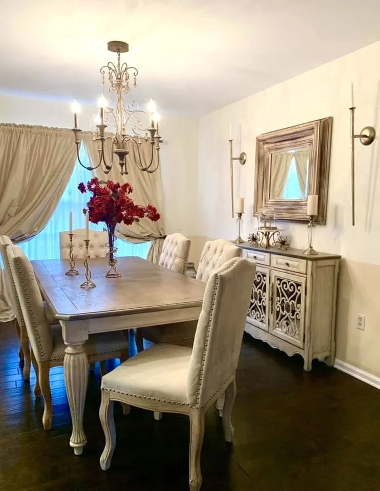Elegant dining room with a wooden table, six upholstered chairs, a chandelier, a sideboard with decorative items, a large mirror, and curtains.