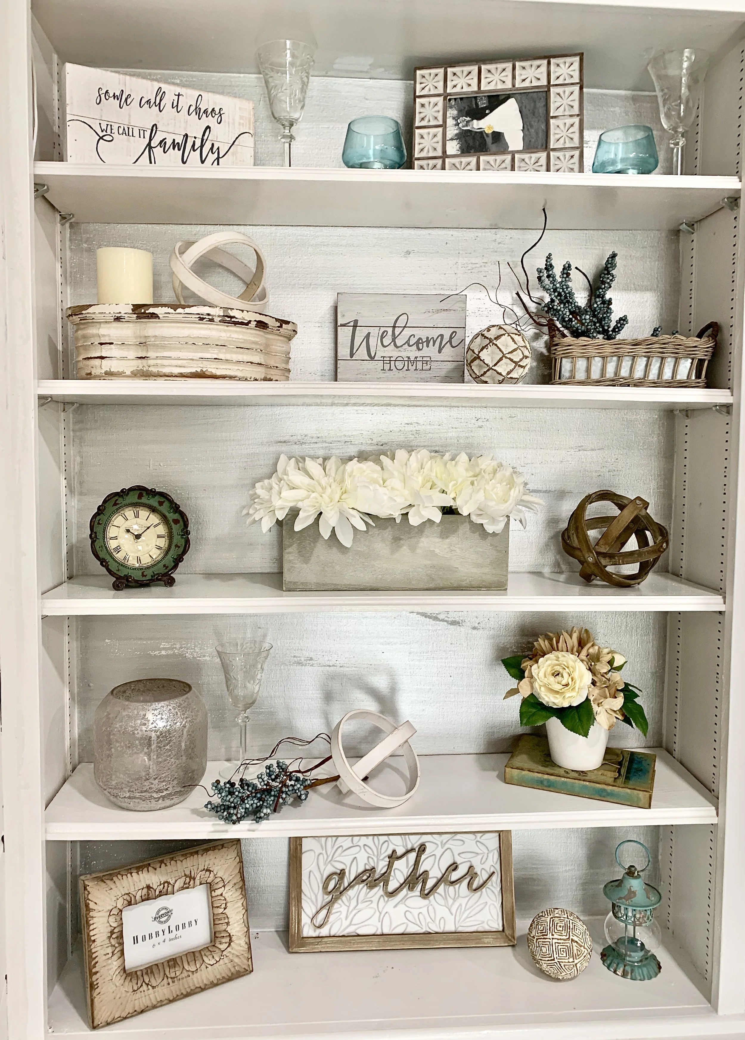 A white bookshelf decorated with various home decor items including flowers, framed signs, glassware, a clock, and decorative objects in neutral and pastel tones.