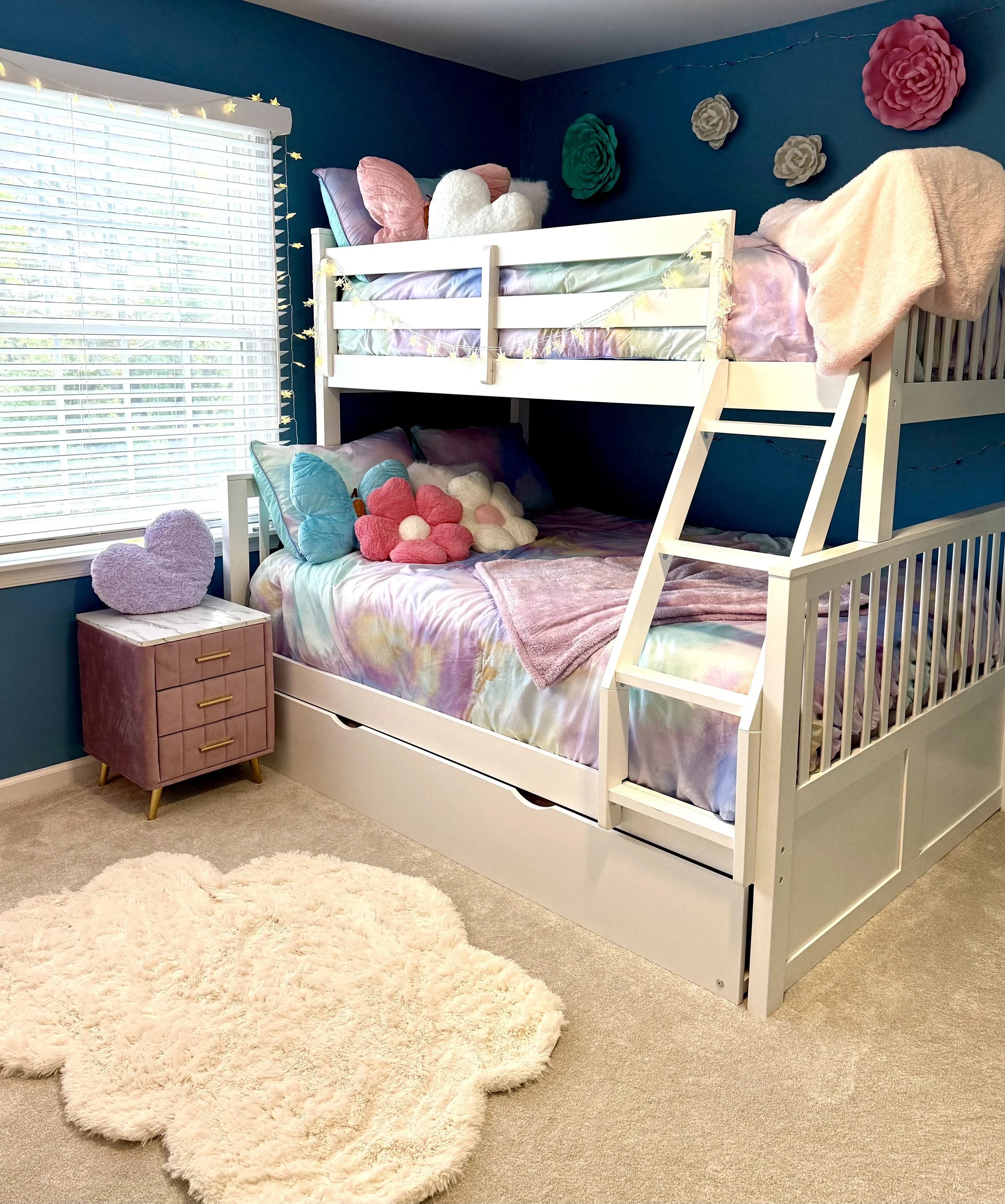A bedroom with a white bunk bed against a dark teal wall decorated with large, colorful flower wall art. The top bunk has pastel bedding and plush pillows, including a heart-shaped pillow, while the bottom bunk has a galaxy-themed bedspread with plus