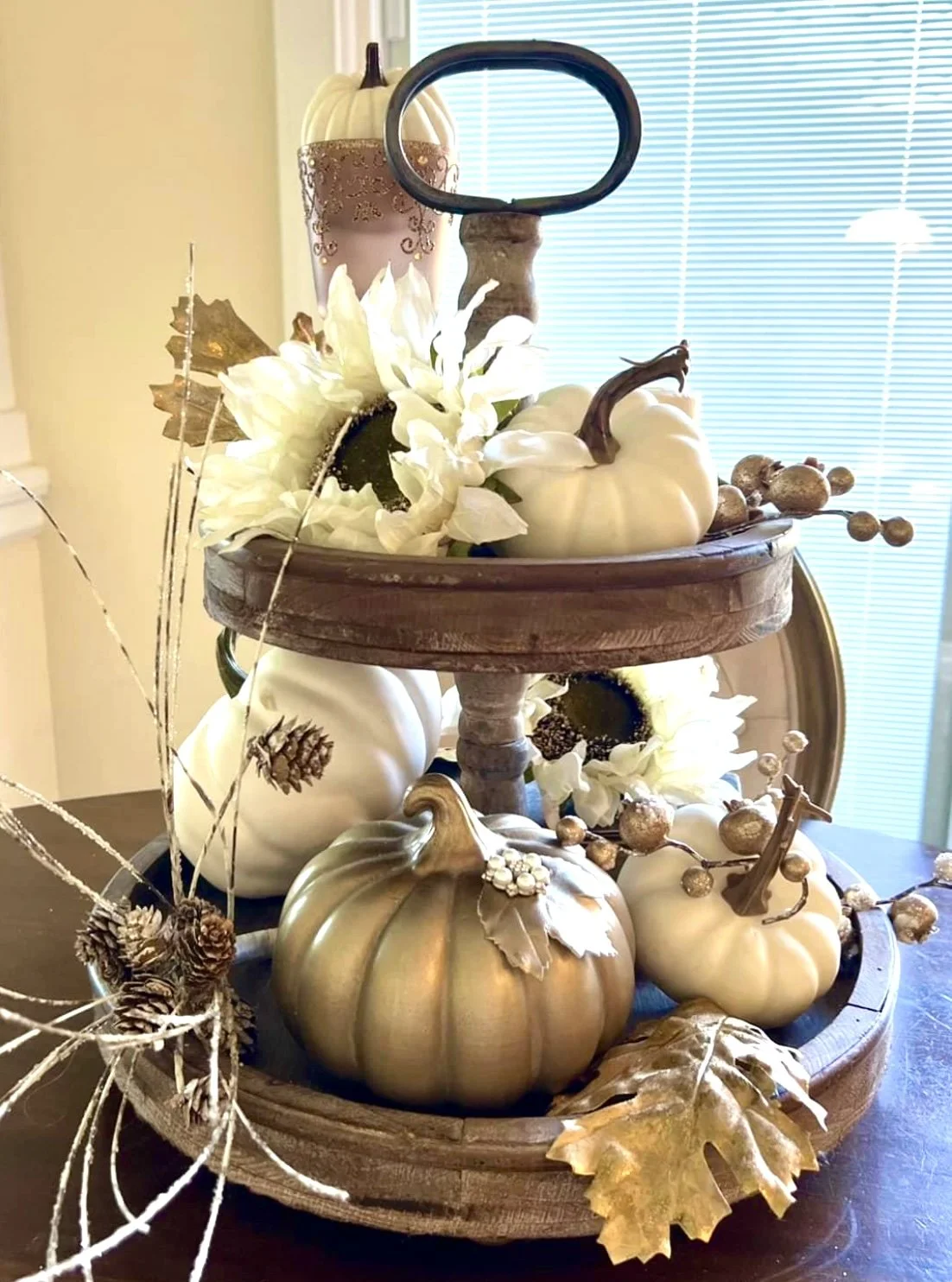 Decorative autumn centerpiece featuring white pumpkins, a silver pumpkin, artificial white flowers, gold-colored leaves, pinecones, and accents, arranged on a two-tiered wooden tray on a table near a window with blinds.