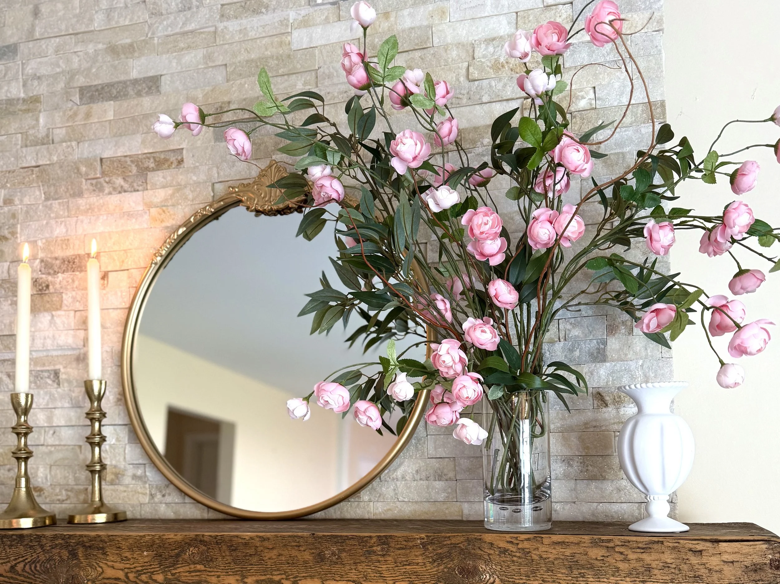 Fireplace mantel styled with a round gold mirror, pink floral arrangement, and soft candlelight against a textured ledger stone surround.
