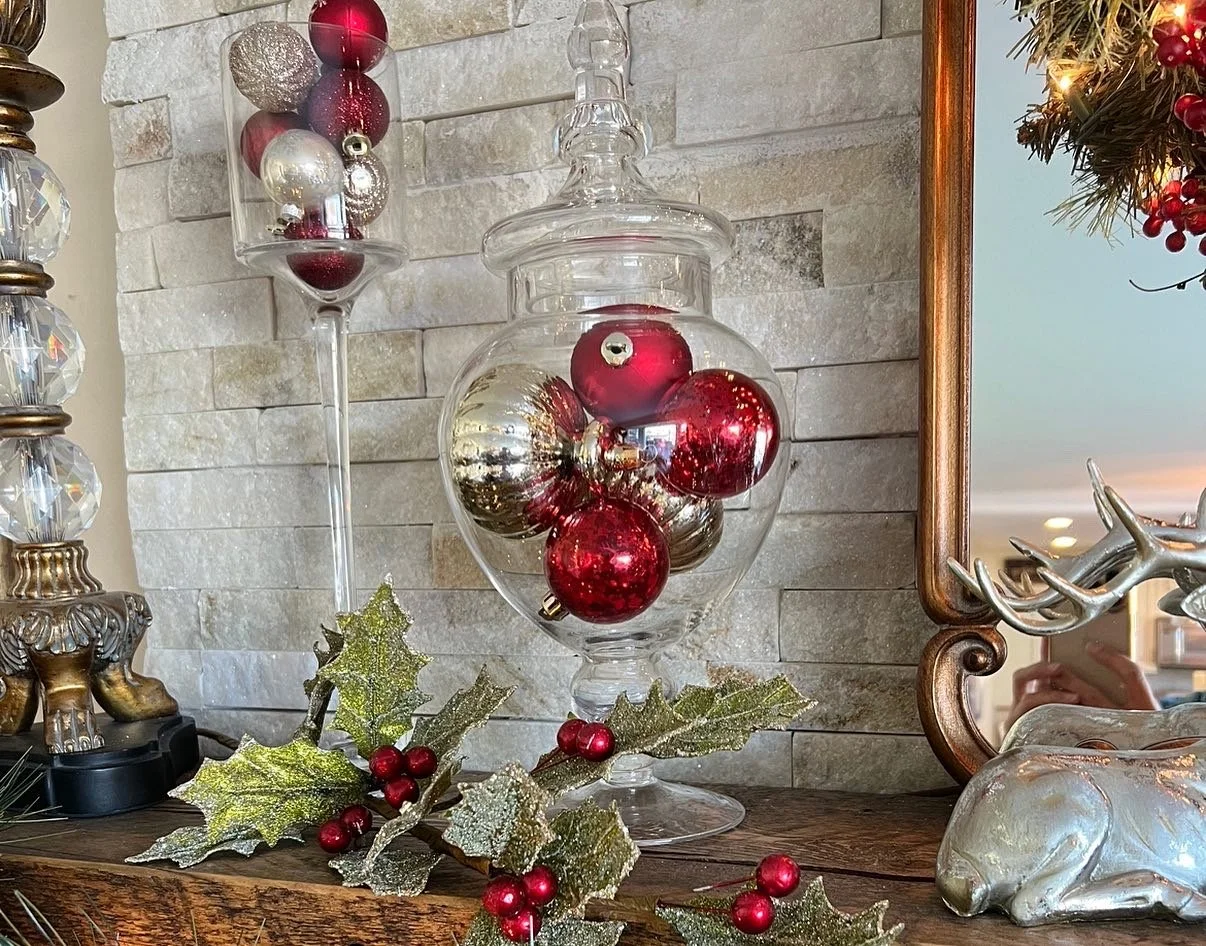 A holiday display with a large glass jar filled with red, gold, and silver Christmas ornaments, a holly branch with red berries, a mirror, and decorative items on a wooden surface against a stone wall.