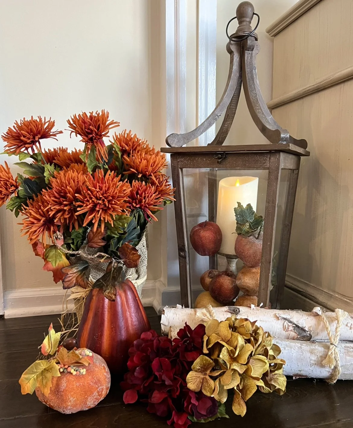 Fall-themed decor with orange flowers, artificial pomegranates, a lit candle inside a lantern, a pumpkin with leaves, and a rolled scroll on a dark wooden surface.