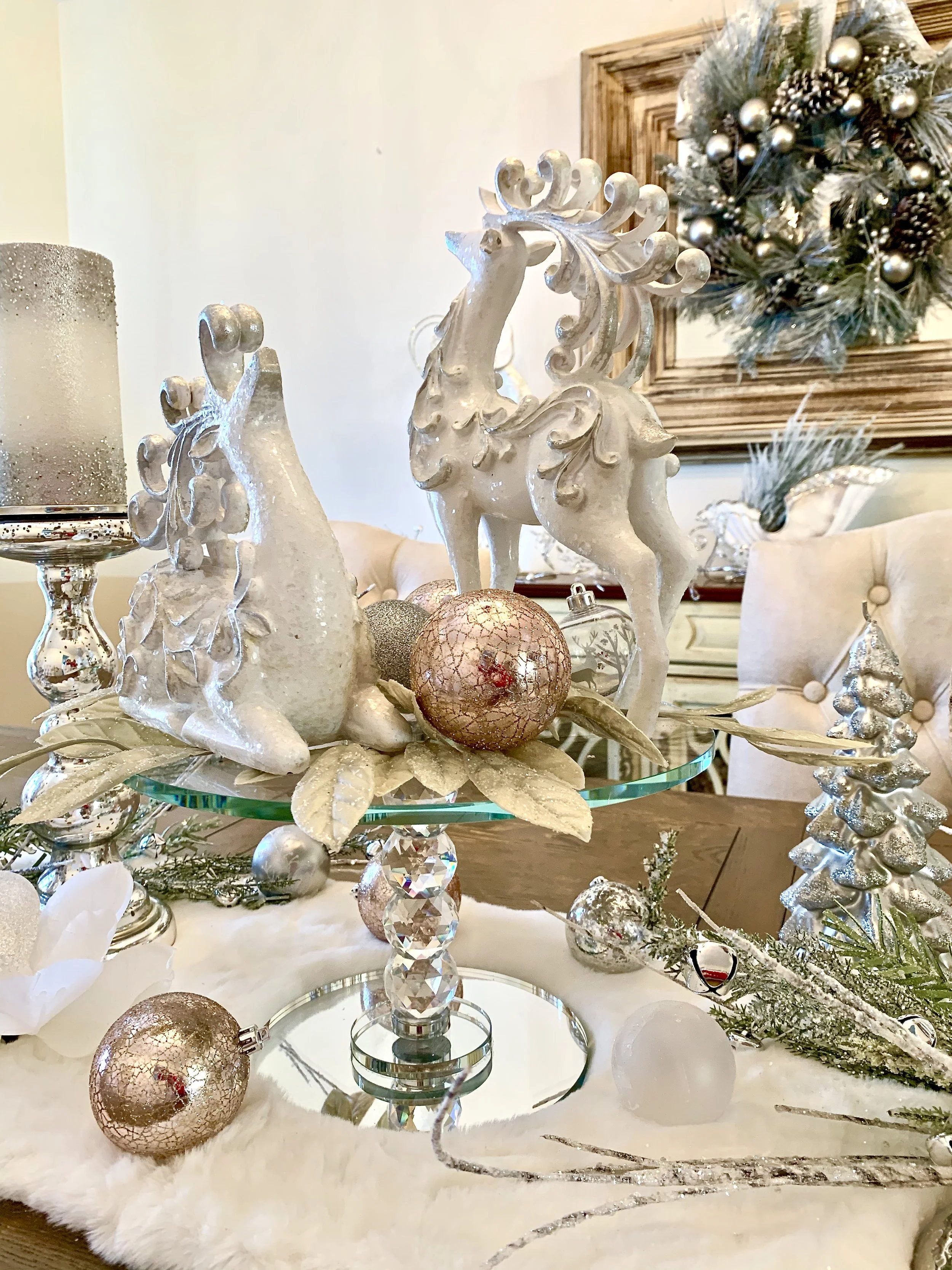 Decorative Christmas centerpiece with white reindeer figurines, glass ornaments, and faux leaves on a mirrored cake stand, surrounded by a plush white surface, with a Christmas wreath hanging on the wall in the background.