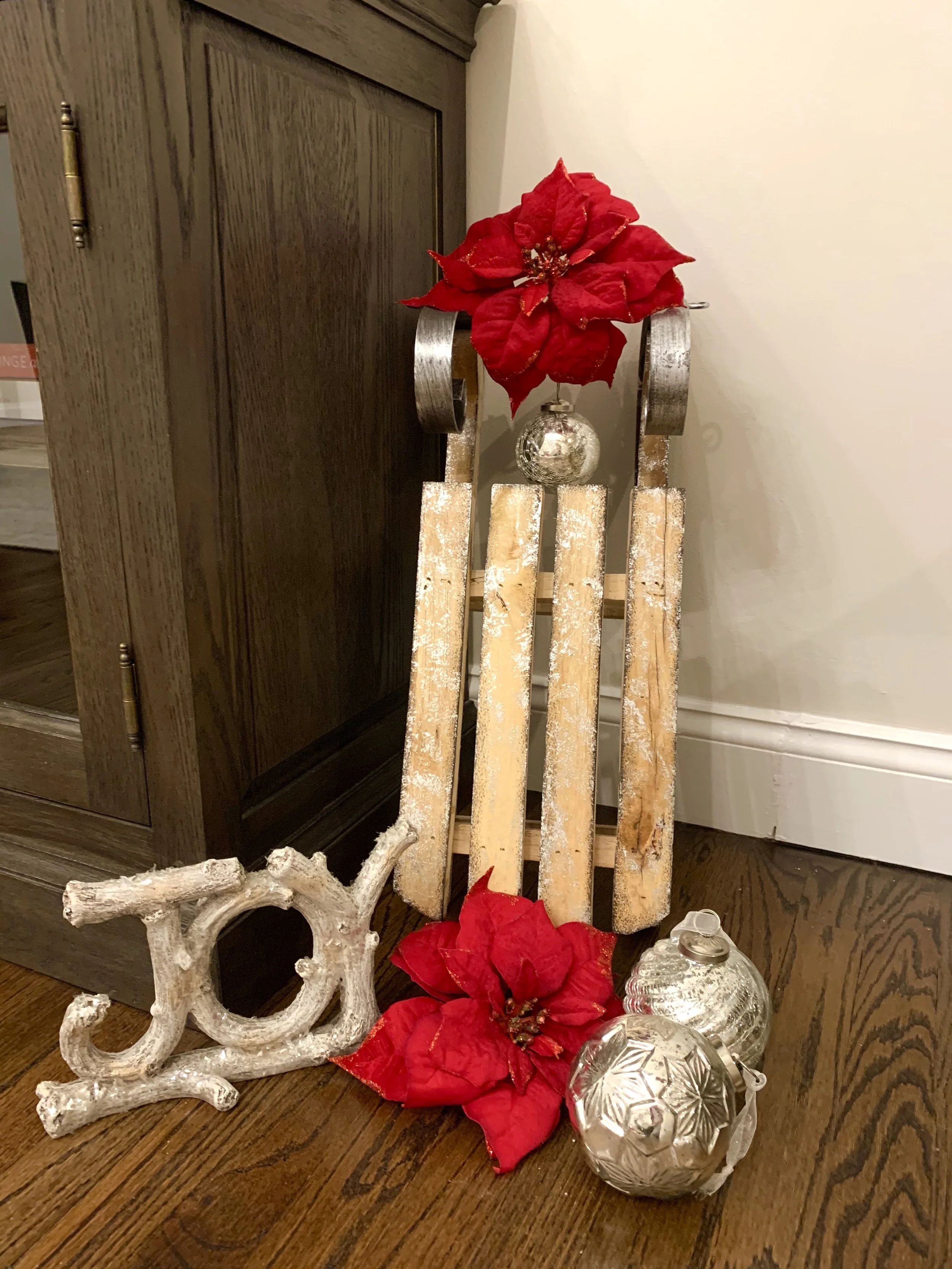 Decorative Christmas display featuring a wooden sled with poinsettia flowers, silver ornaments, and a white driftwood piece, arranged on a hardwood floor near a wooden cabinet.