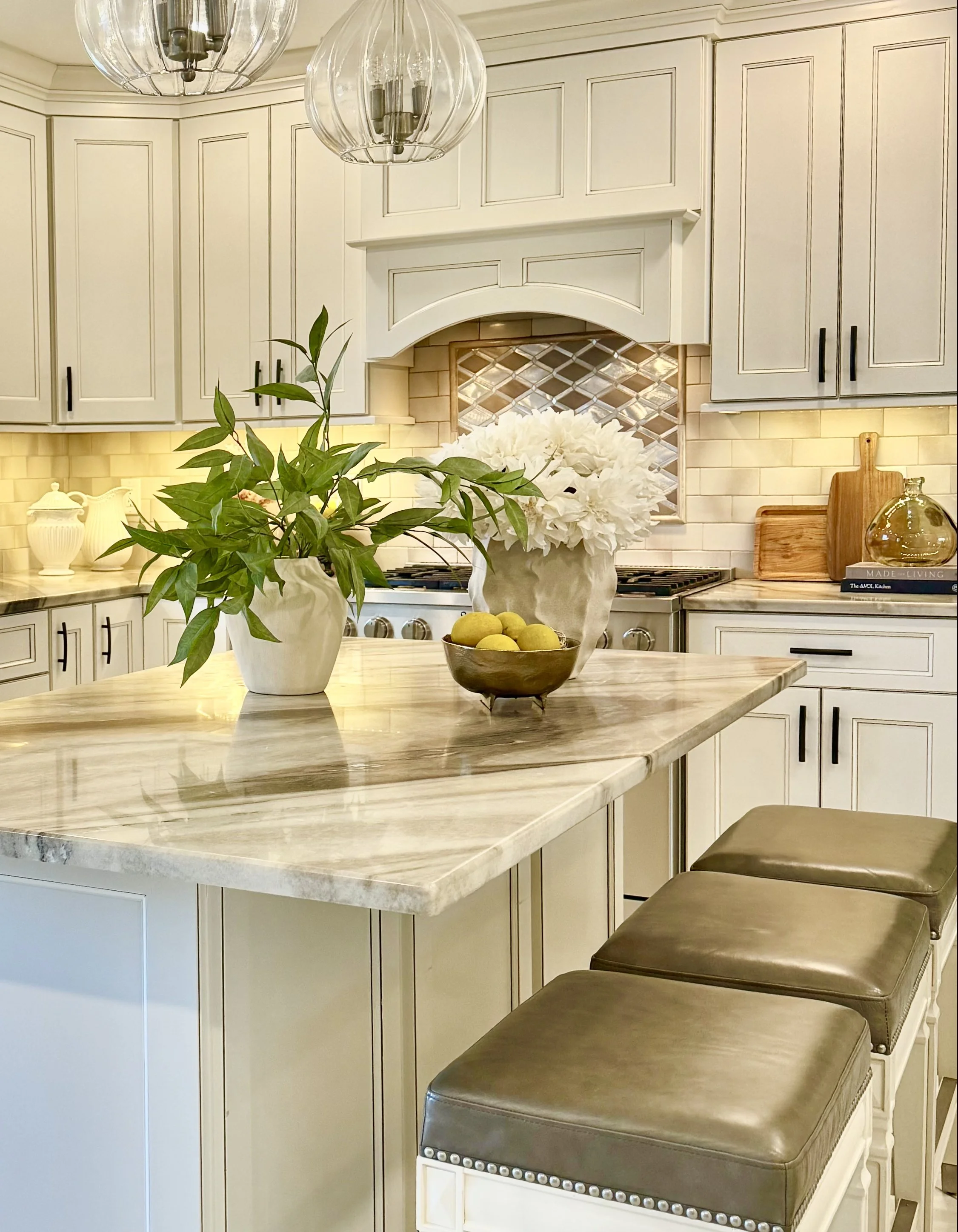 Elegant white kitchen with enlarged countertops, upholstered bar stools, glass pendant lighting, custom cabinetry, decorative greenery, beige, brown and white with black pulls. Designed for a warm and inviting luxury home interior. 