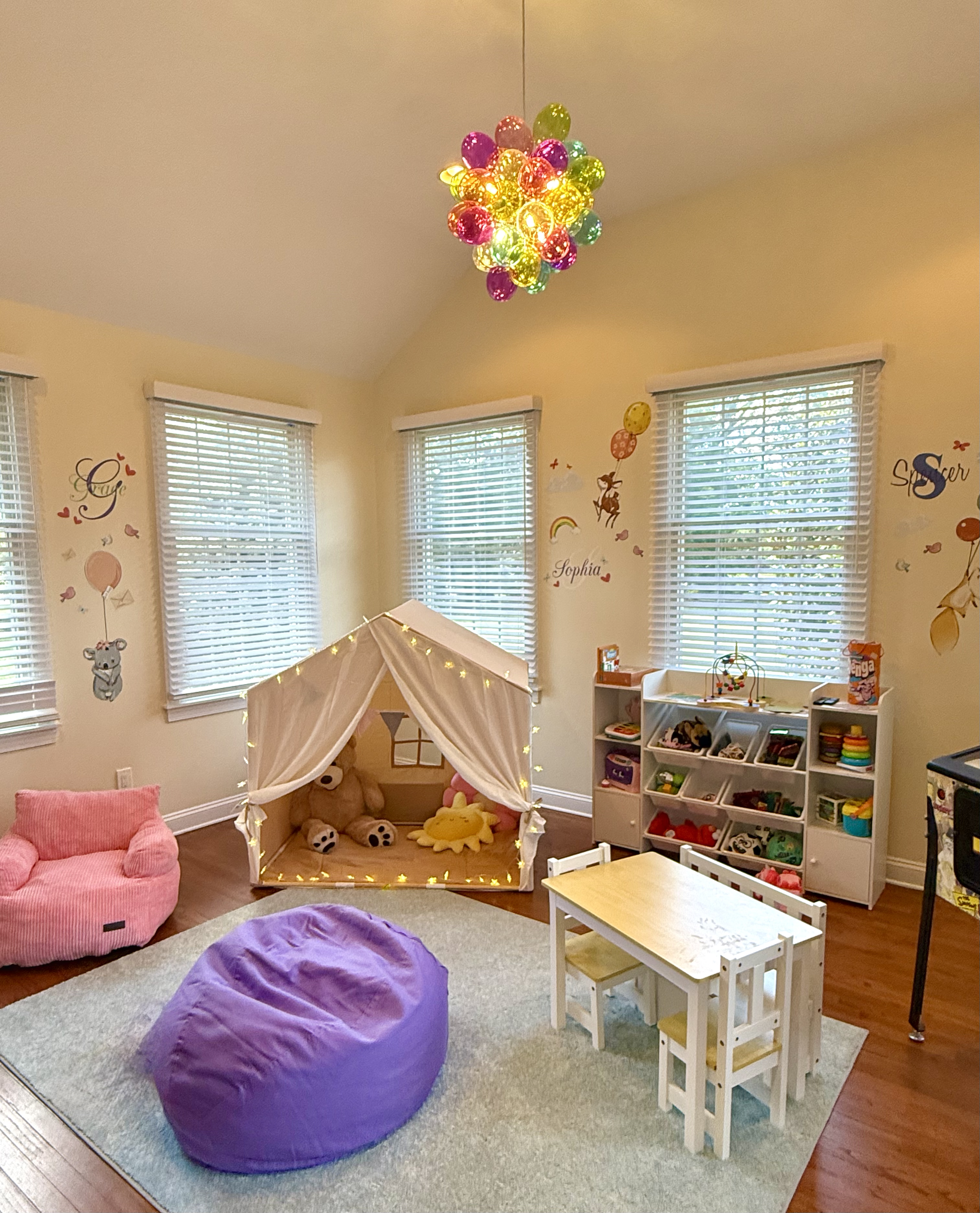 Children's playroom with a teepee tent, stuffed animals, pink couch, purple bean bag, small table with chairs, storage shelves with toys, colorful balloon chandelier, and wall decals in the background.