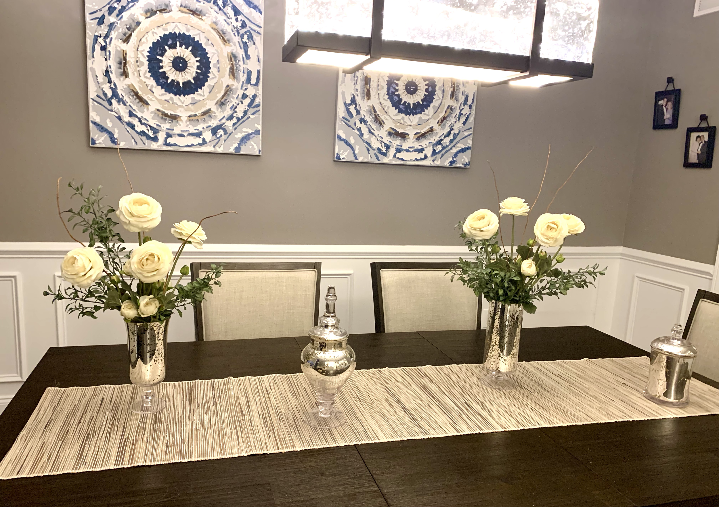 A dining table with a beige table runner, two silver vases with white roses and greenery, and two decorative silver urns, in a room with gray walls, white wainscoting, and two abstract blue and white paintings.