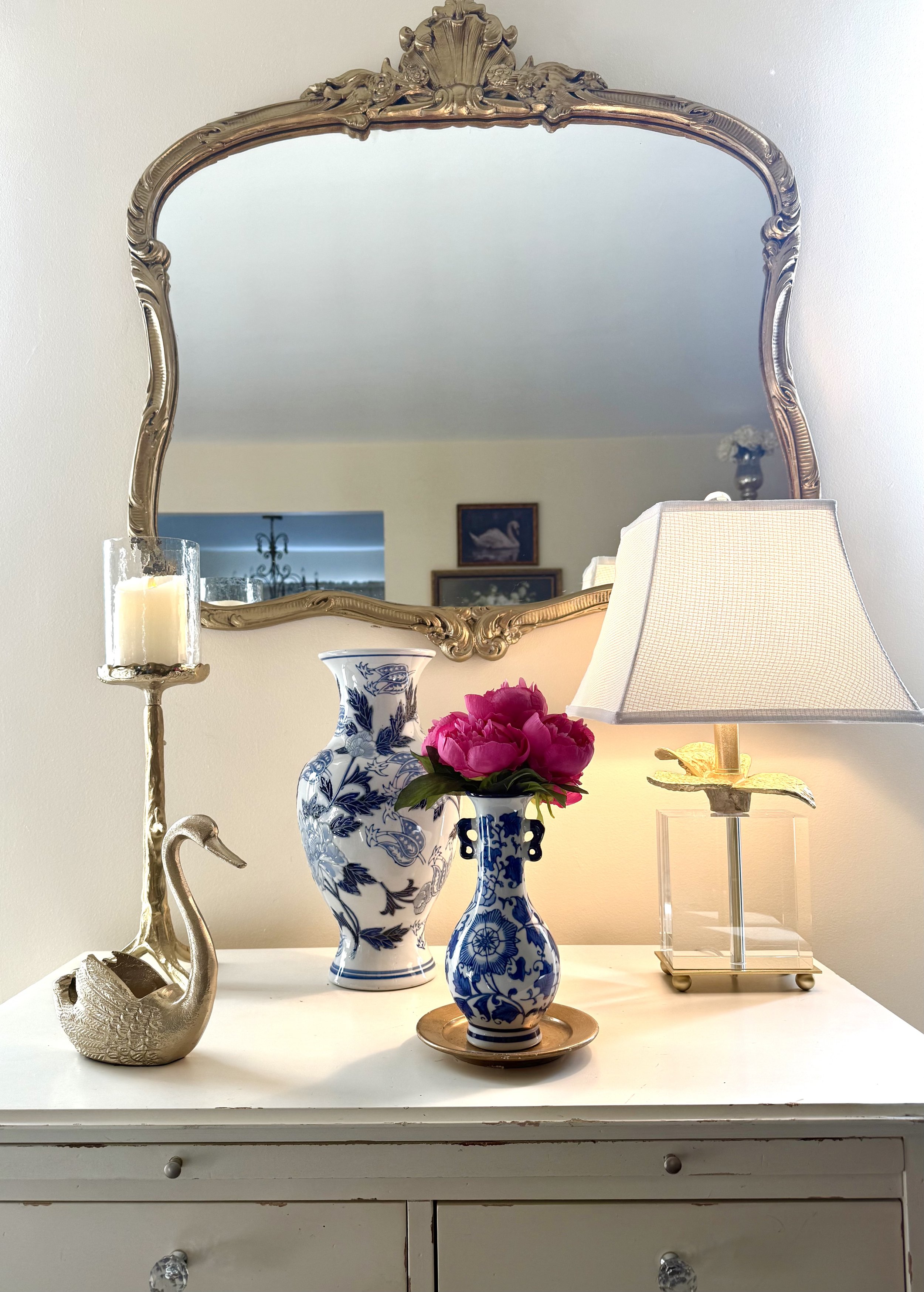 This vignette features a vintage dresser styled as an elegant focal point, anchored by an ornate gold mirror that adds height and architectural interest. The crystal and gold table lamp adds warmth while blue and white porcelain accents introduce cla