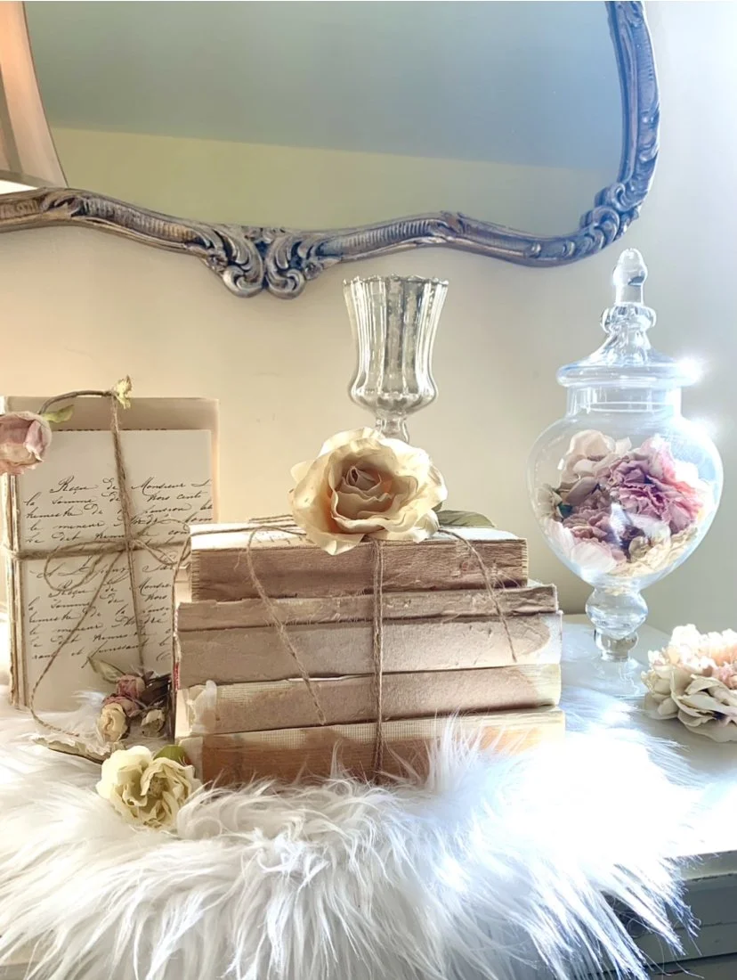 Decorative vintage scene with books tied with twine, fresh and dried flowers, a glass vase with a flower, a glass jar with flower petals, all on a white fluffy surface, with an ornate mirror above.