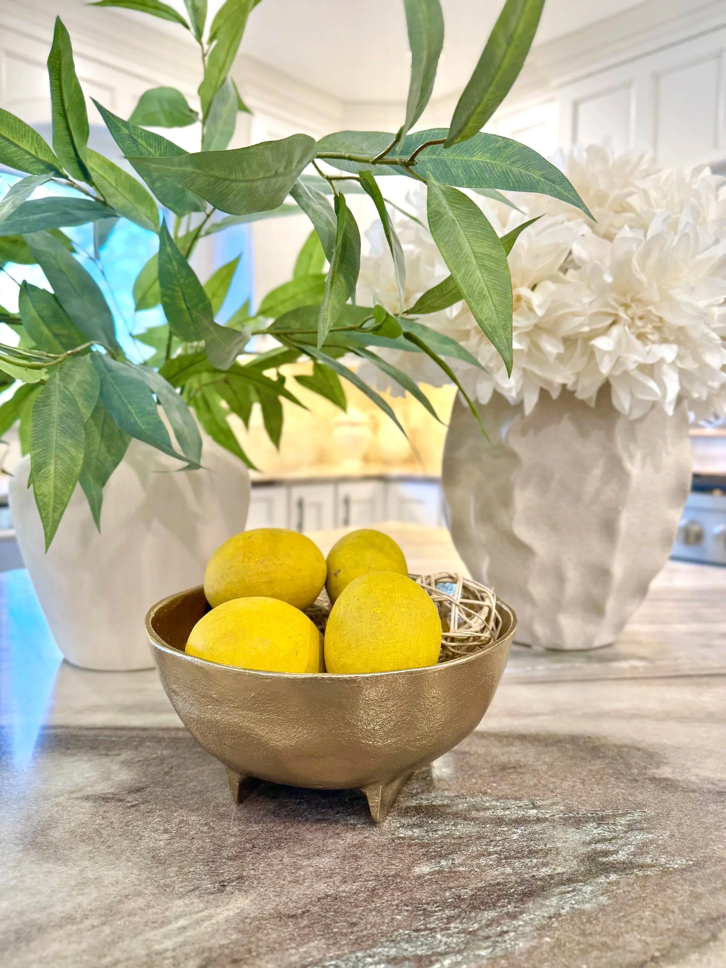A gold bowl filled with lemons on a kitchen island brown and tan countertop layered with a white vase filled with greenery and white flowers.