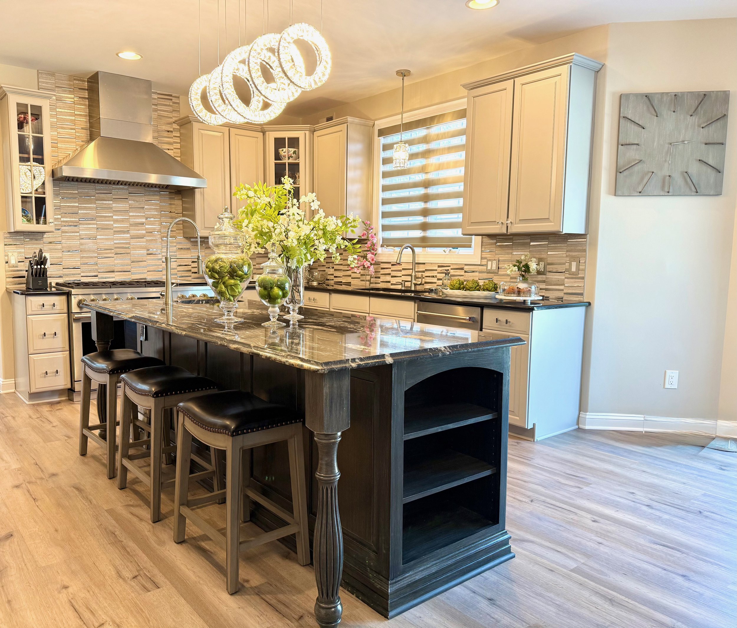 Remodeled kitchen with grey cabinets, a black island, light wooden floors, a modern crystal chandelier, wooden stools with dark grey leather seats, a tiled tan and mirrored backsplash styled with a large green and white flower arrangement in a glass 