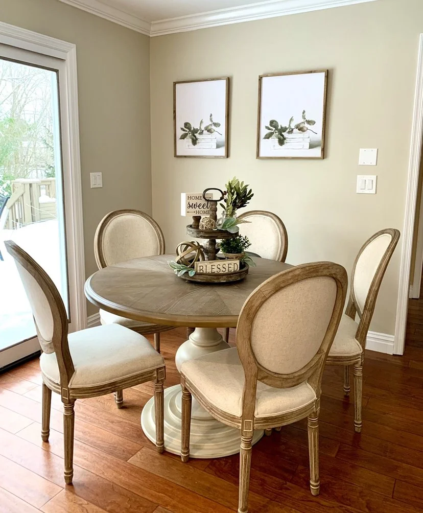 A round wooden dining table with five upholstered cream-colored chairs in a dining room. Centered on the table is a decorative tray with plants, a sign that reads 'Home Sweet Home', and a small wooden block with the word 'Blessed'. On the wall behind