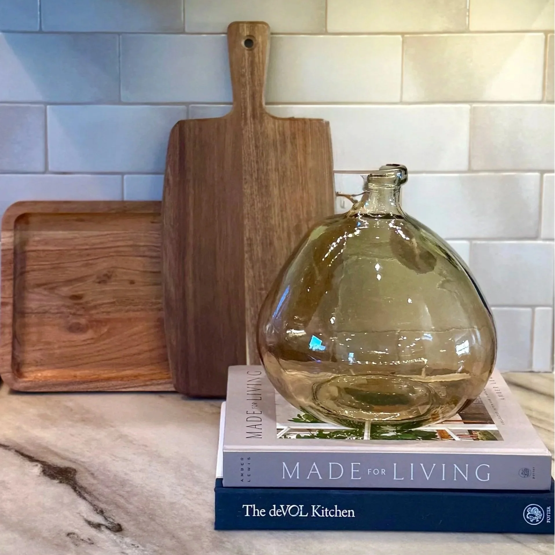 Layered cutting boards leaning on a glass tan kitchen backsplash with decorative books in front with a clear brown vase sitting on top.