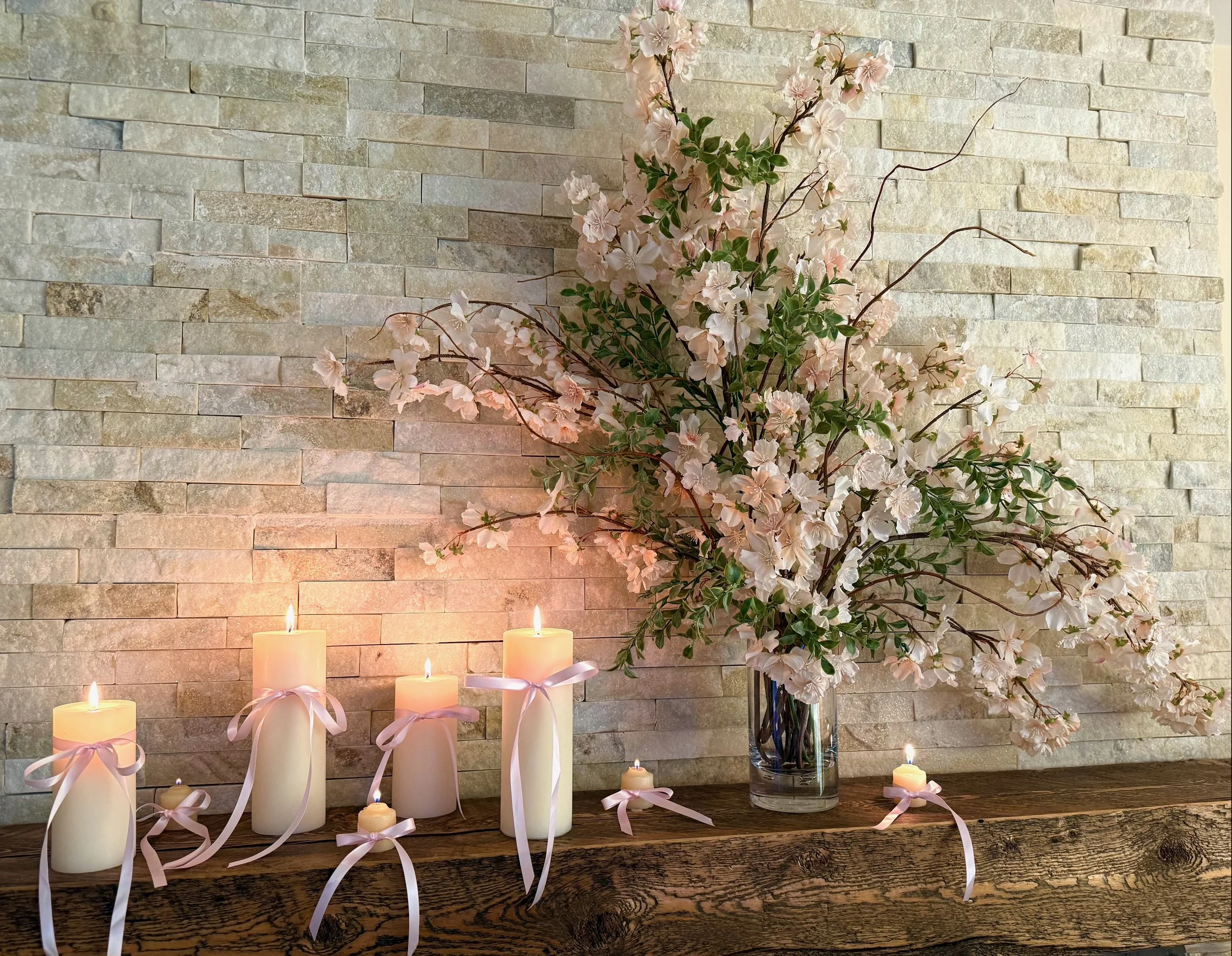 fireplace with rustic wooden mantle, beige and white ledger stone surrounding with a cherry blossom floral arrangment and cream candles of various heights tied with a pink ribbon