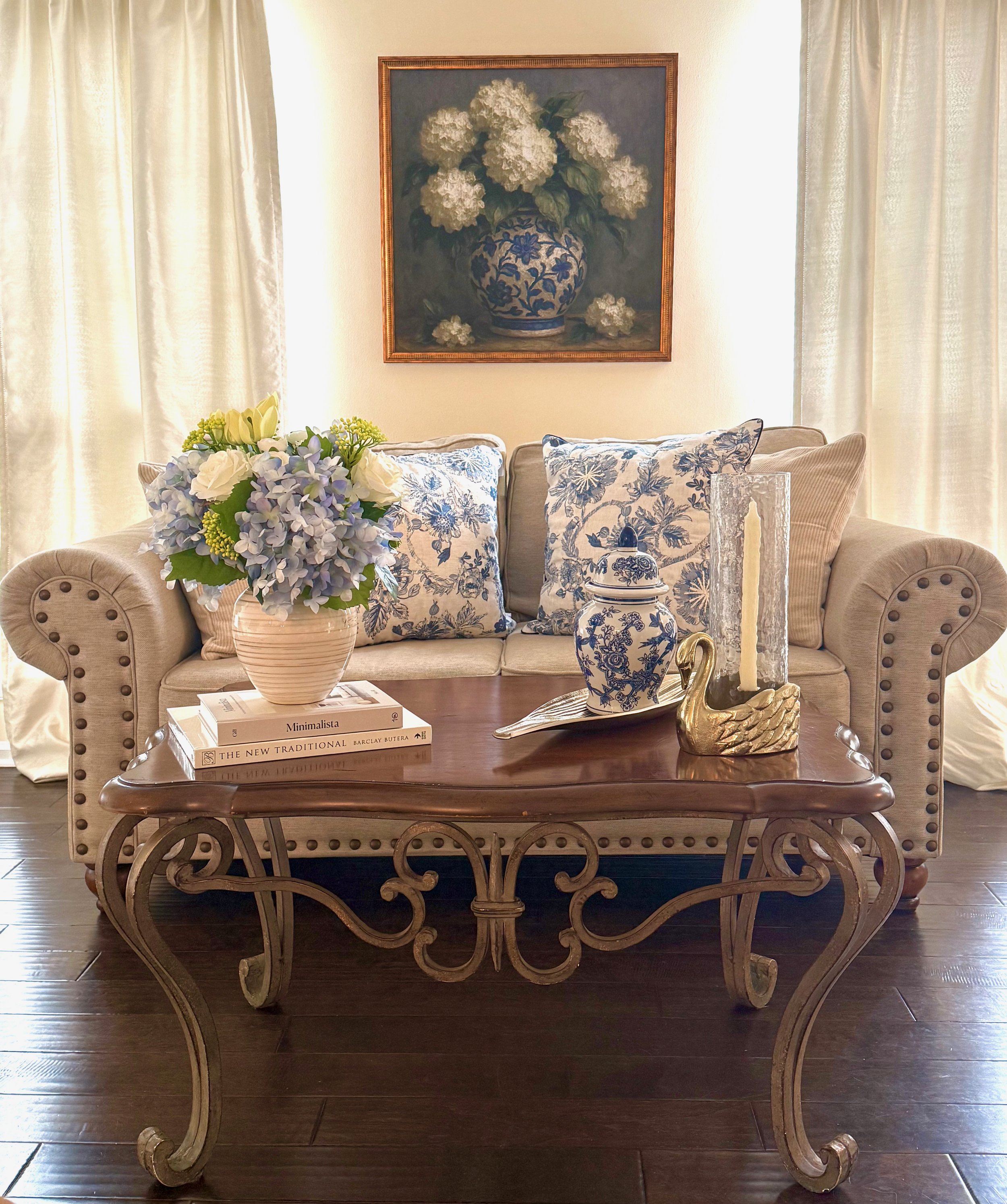 Professionally classic styled living room with a neutral beige sofa, blue and white patterned pillows, silk drapery, gold framed blue and white artwork, layered textiles, an ornate iron and wood coffee table, dark wood floors, gold swan hurricane can