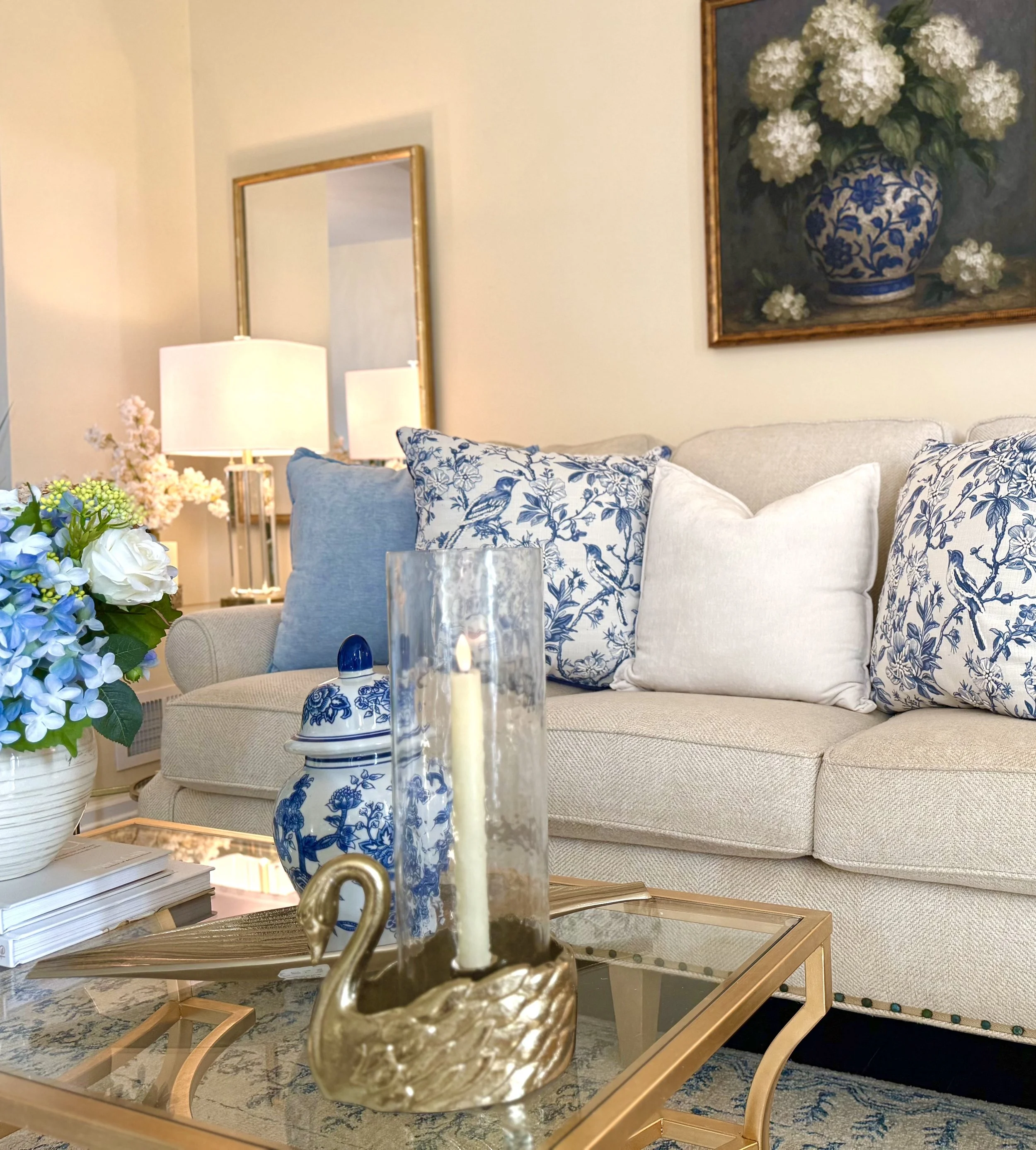 A thoughtfully styled living room featuring a neutral sofa layered with blue and white patterned and solid pillows, flanked by matching crystal and gold table lamps on gold and glass end tables and gold mirrors behind each lamp. Gold framed artwork s