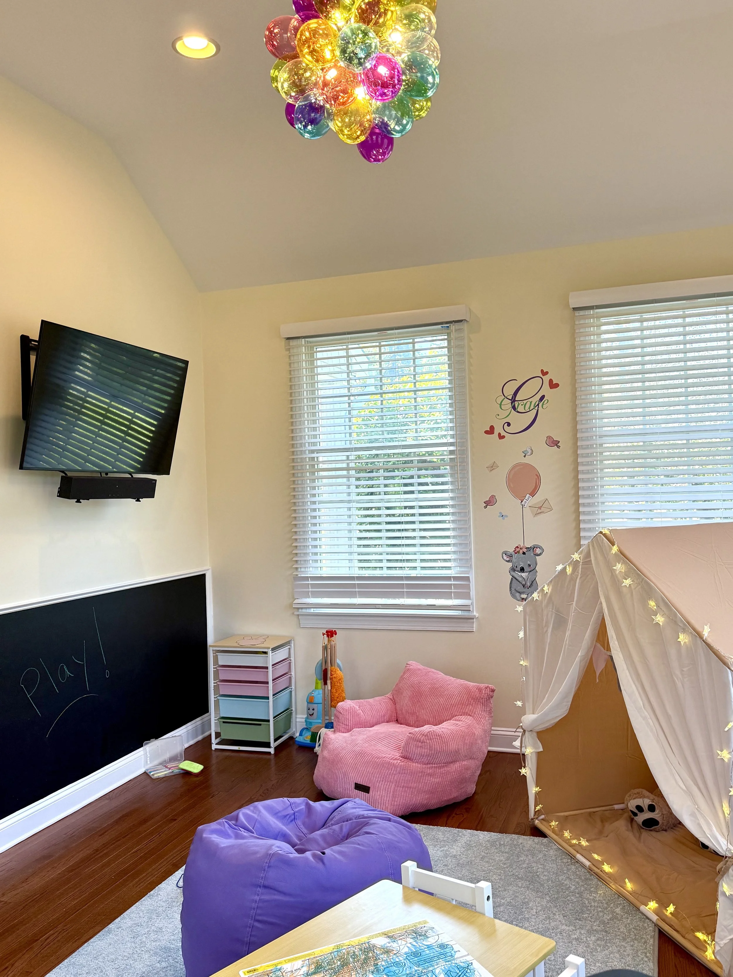 Colorful glass ball chandelier hanging from a ceiling in a children's playroom with pink and purple seating, a play tent with lights, a chalkboard, and wall decorations.