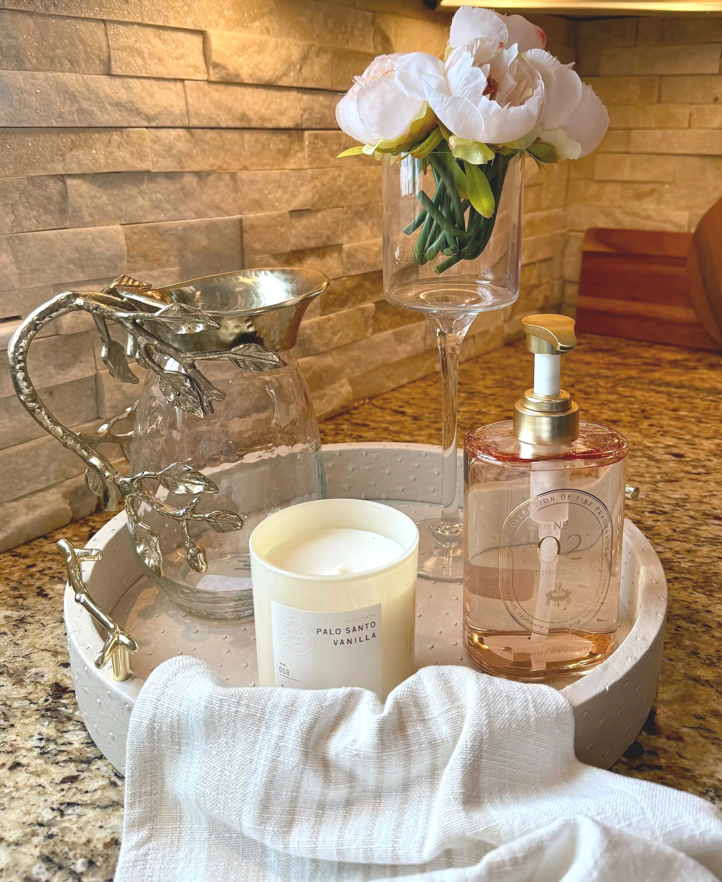 beautiful kitchen beige and gold tray styled with a glass and gold pitcher, a candle, gold and pink soap dispenser and glass fluted cup holding pink peonies with a dish towel with cutting boards against a cream and brown ledger stone backsplash