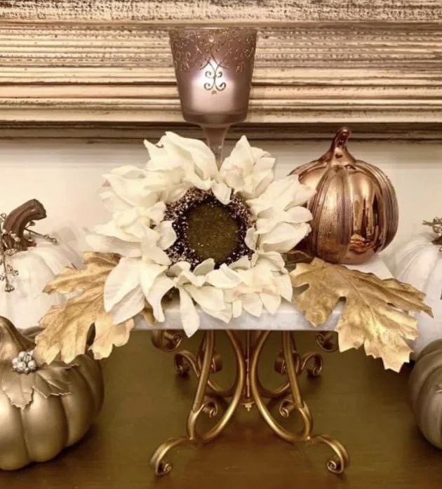 Decorative fall centerpiece featuring artificial pumpkins in metallic bronze, white flowers, gold leaves, a purple candle in a decorative holder, all arranged on a table.