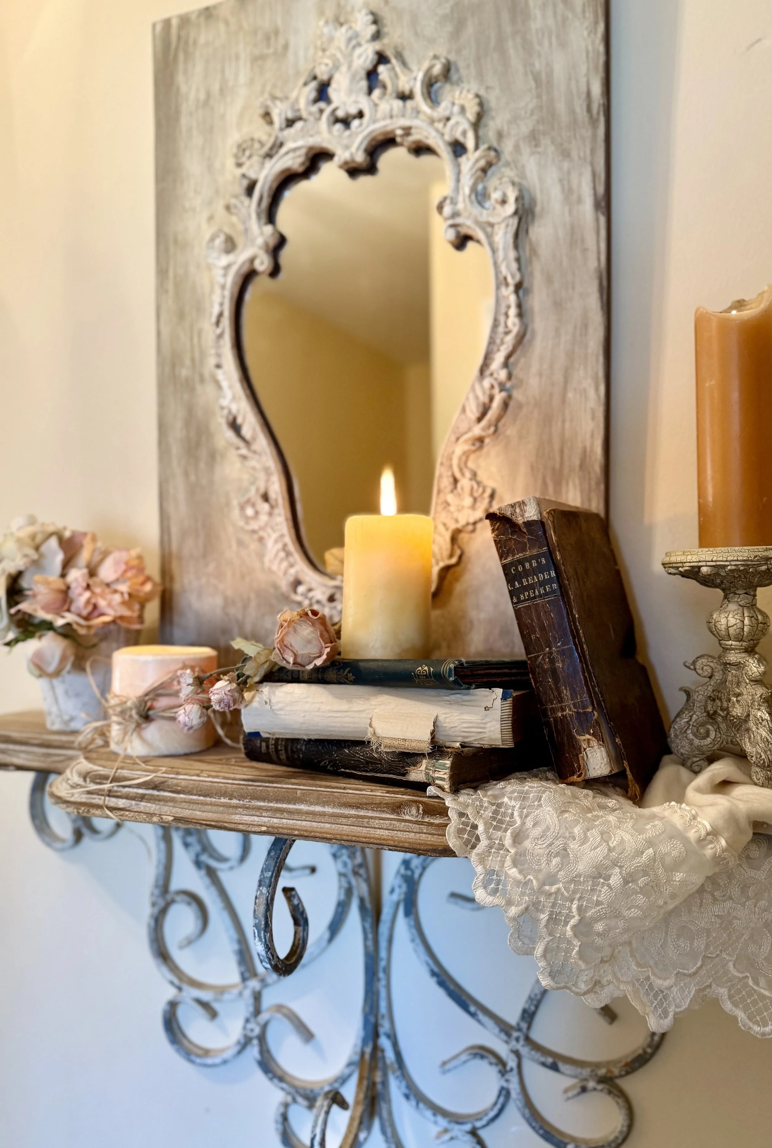 Decorative wall shelf with ornate metal scroll with a rustic ornate mirror, soft pink flowers, old worn books with a candle placed on top, lace fabric hanging
