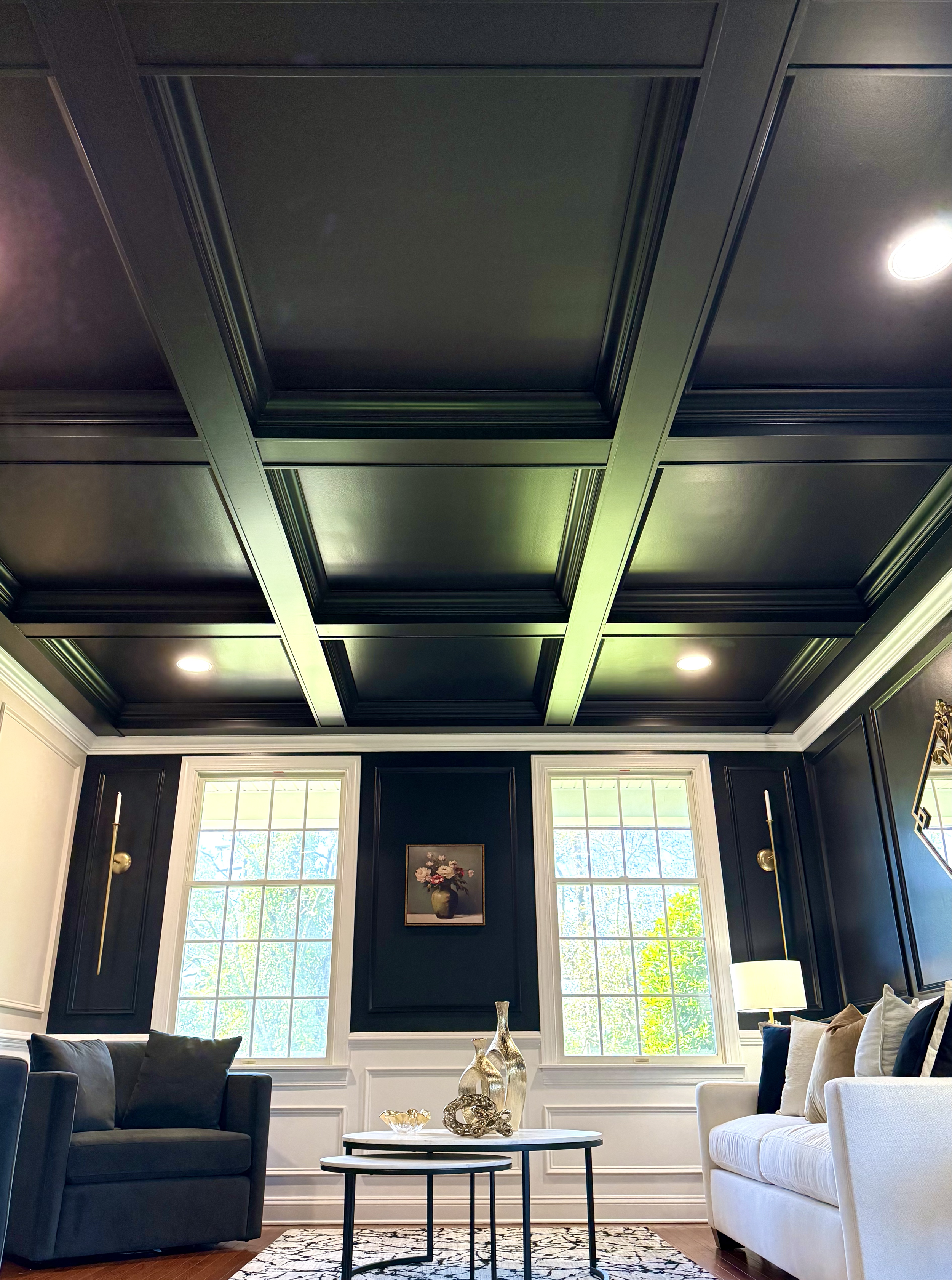 Living room with dark coffered ceiling, two windows, black and white walls, couches, coffee table with decorative vases and sculptures, and a painting on the wall.