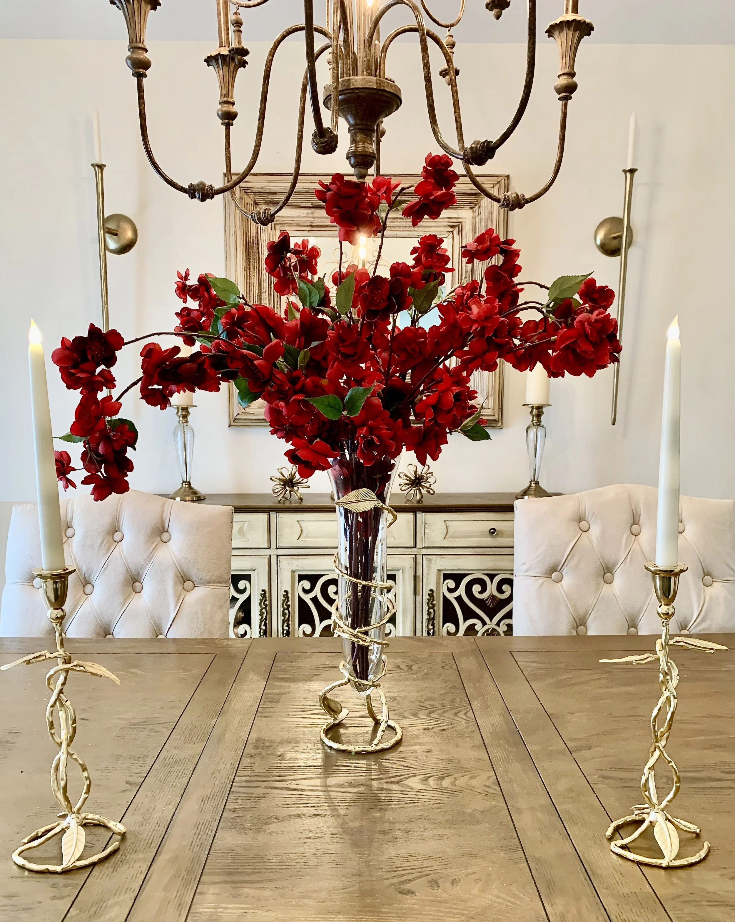 A dining room table with a tall glass vase of red flowers, surrounded by gold candlesticks with white candles, and a chandelier hanging above.