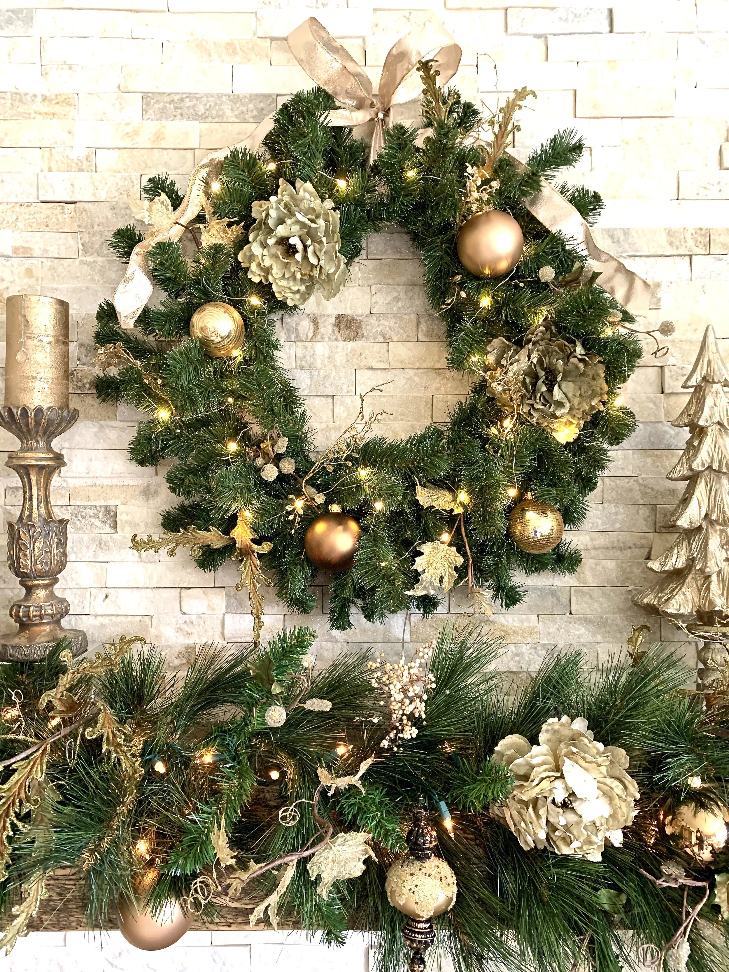 Decorated Christmas wreath with gold and beige ornaments, ribbons, and lights on a stone wall.