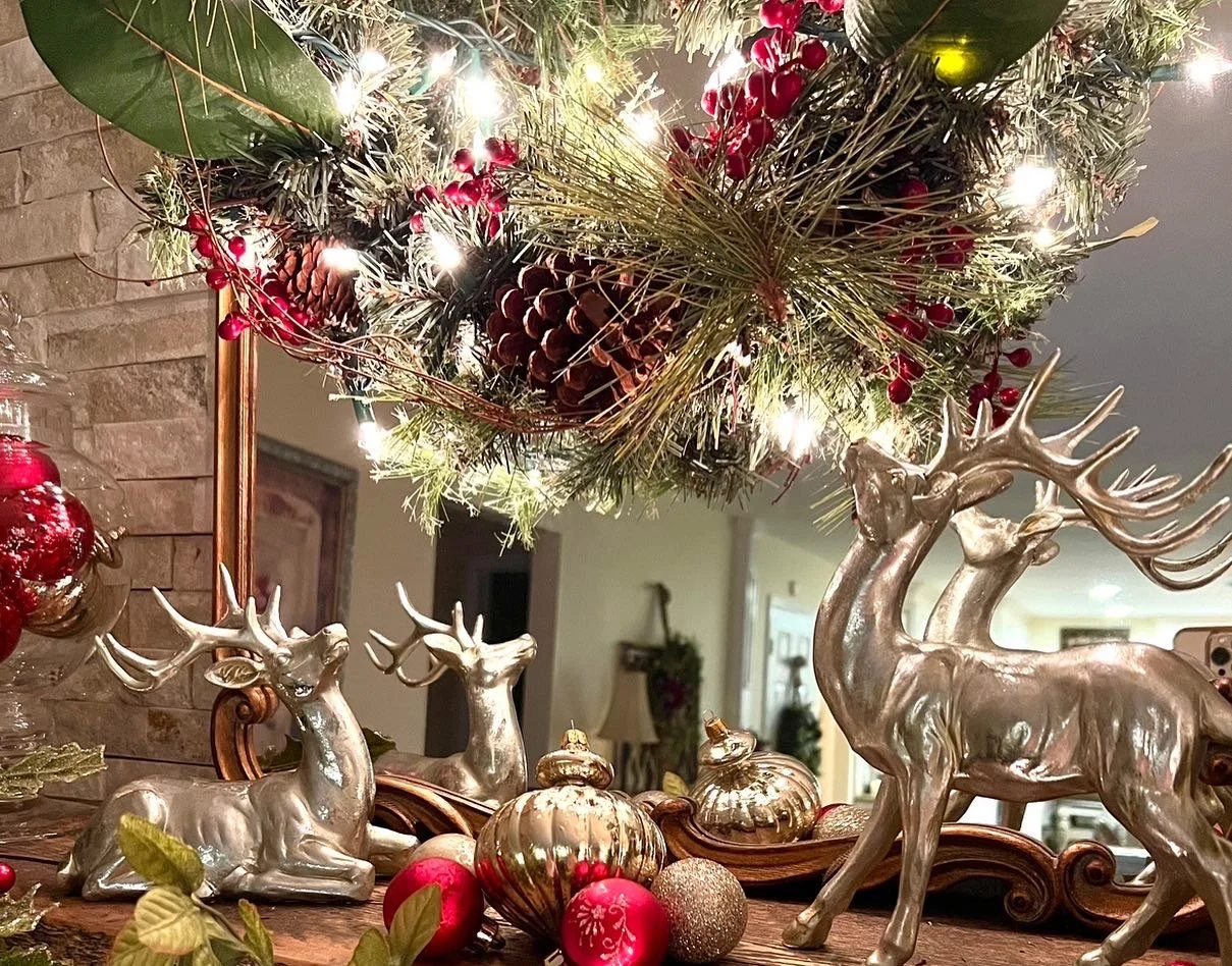 Decorative Christmas arrangement with silver reindeer figurines, red and gold ornaments, pinecones, and greenery with lights, set on a wooden surface near a brick fireplace.