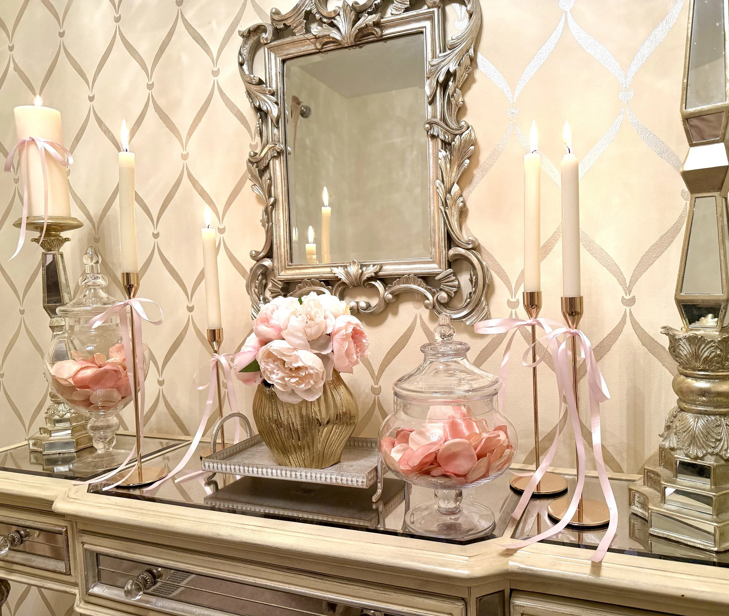 Valentine's Day console decor with soft pink florals in a gold vase, rose petals in clear glass jars, various heights of gold candlesticks tied with pink bows with an ornate silver mirror above.