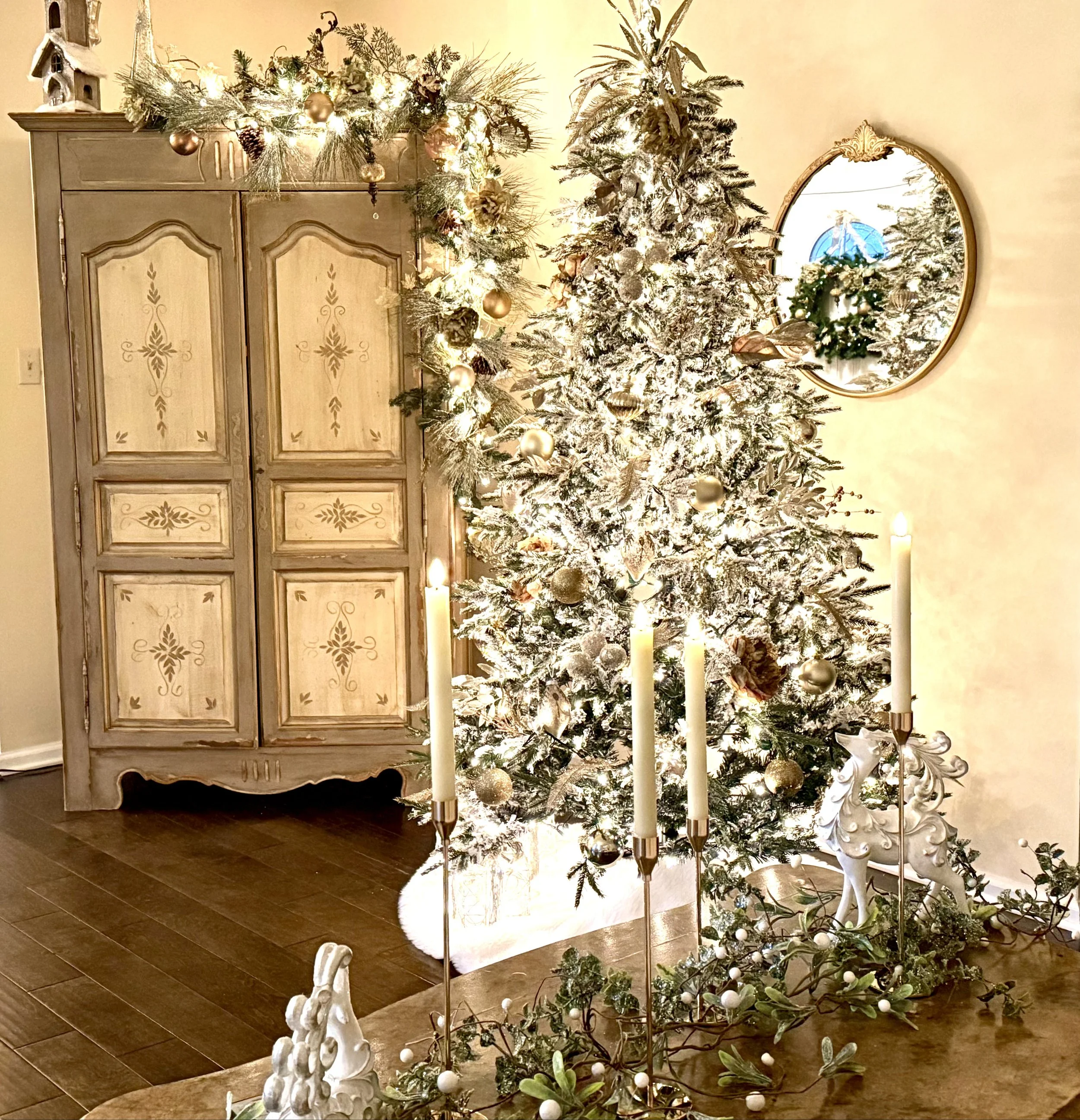 Decorated Christmas tree with gold and white ornaments and a snowy effect, decorated for Christmas with lights. An antique-style white reindeer figurine and candles are arranged on a table nearby. A vintage wooden cabinet with floral patterns and gar