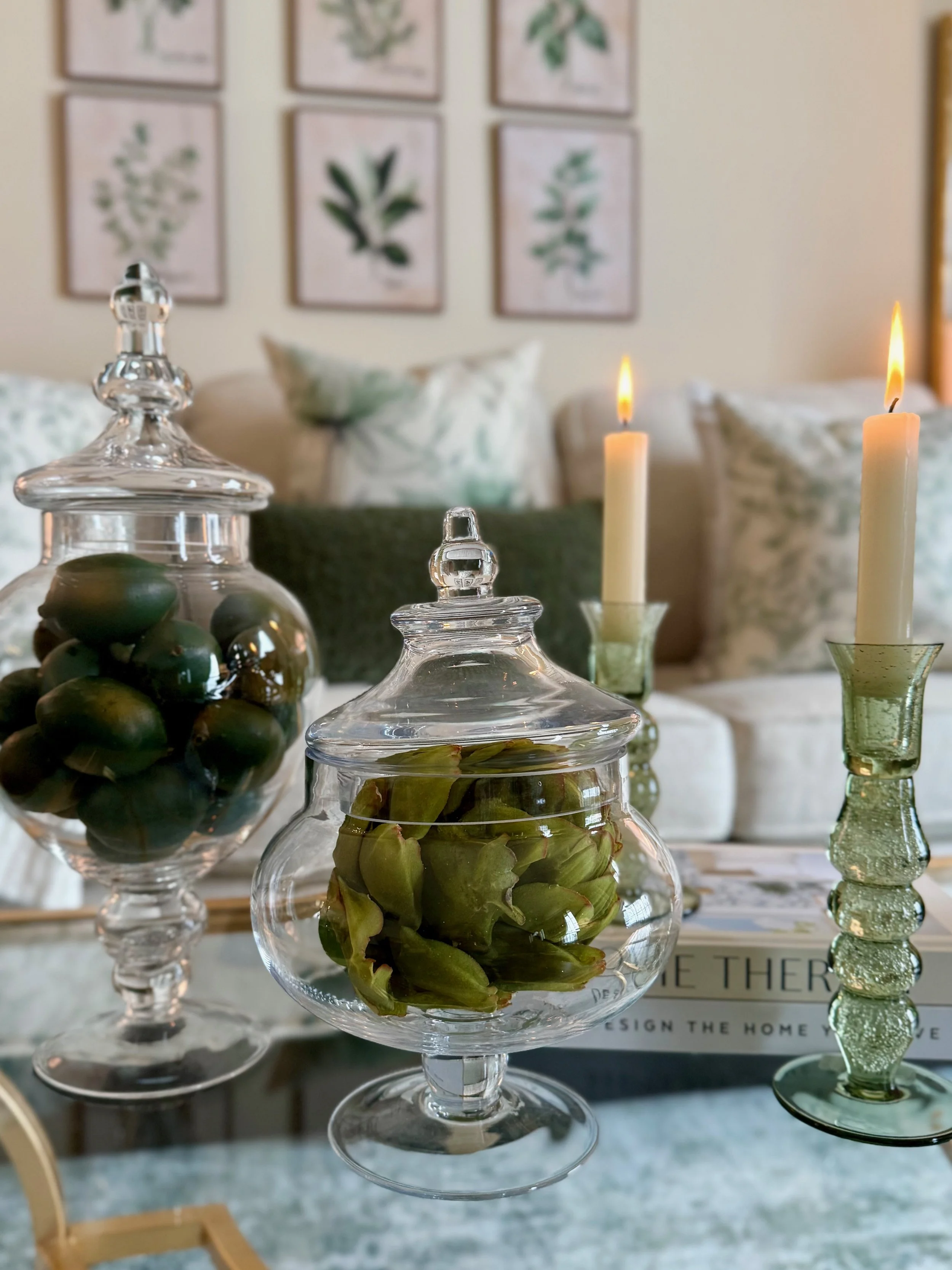 These apothecary glass jars hold green limes and artichokes to accessorize this coffee table in this sage green and white living room along with the green glass candle holders on top of decorative books.