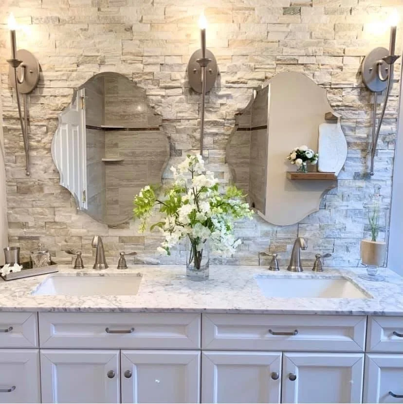 Bathroom vanity with two sinks, marble countertop, two mirrors, and wall-mounted light fixtures with a stone wall background, decorated with a flower arrangement and small decor items.